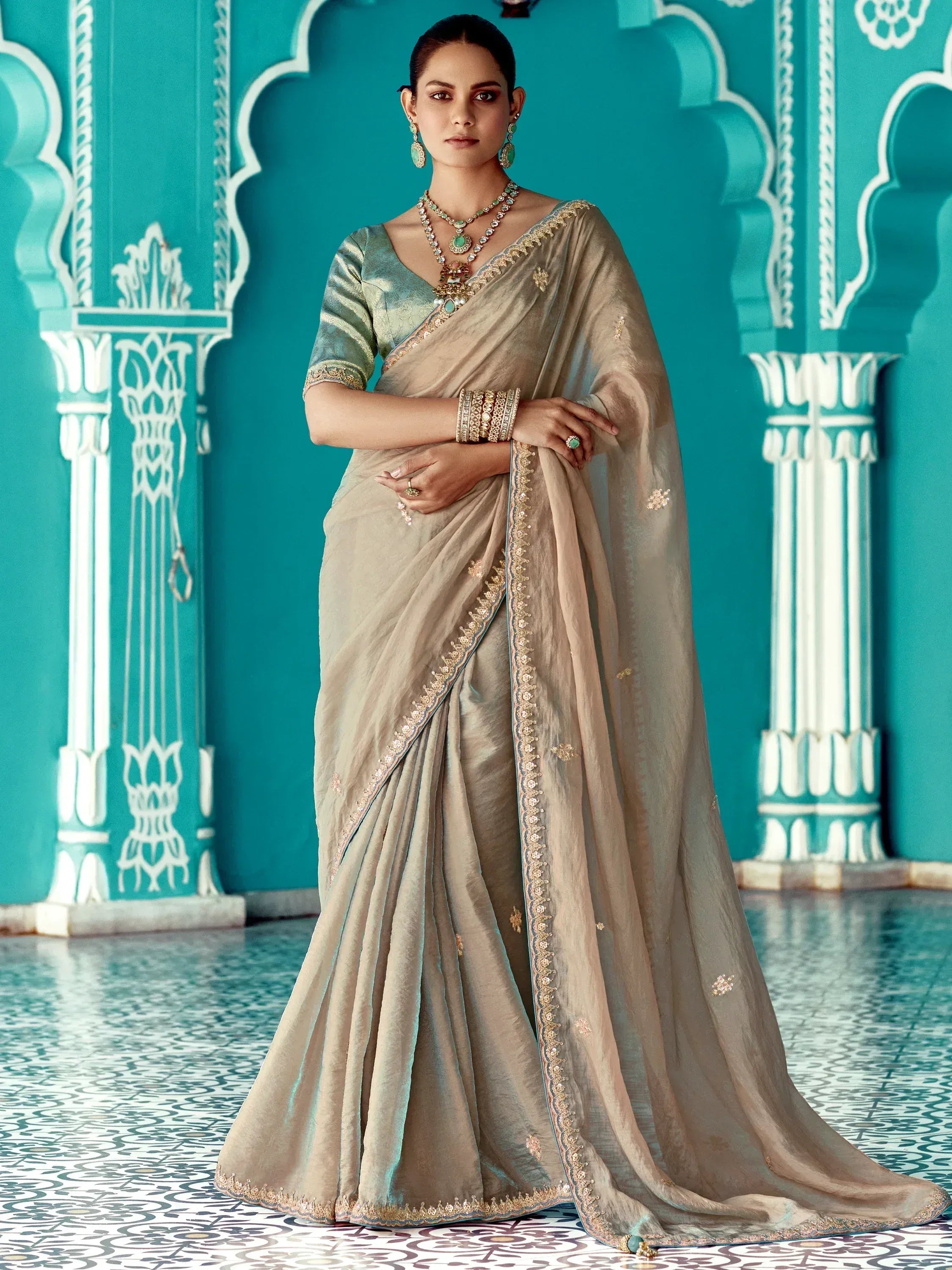 Elegant beige saree with gold embroidery and teal blouse, styled with traditional Indian jewelry against ornate turquoise backdrop