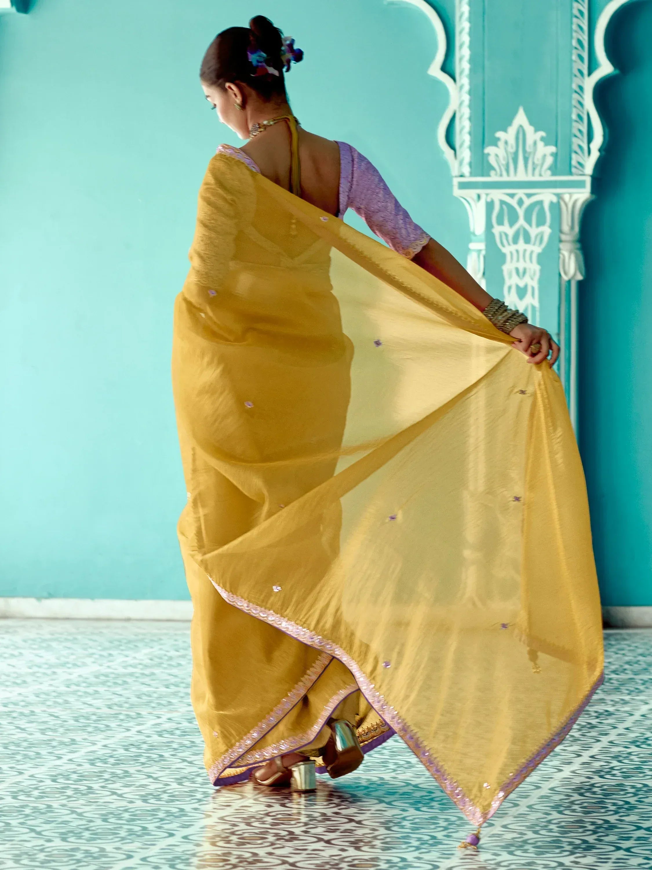 Woman in yellow saree with purple blouse, showcasing ethnic Indian wear against turquoise backdrop
