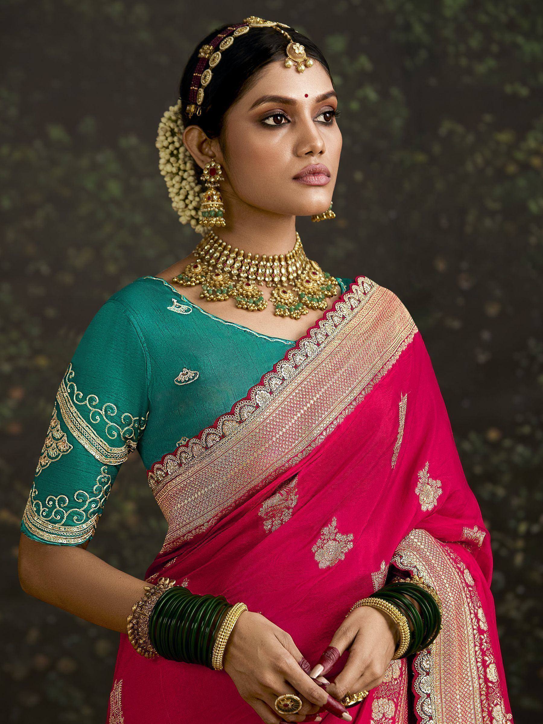 Woman in vibrant pink and green designer saree with gold embroidery, traditional Indian jewelry, and floral hair accessories