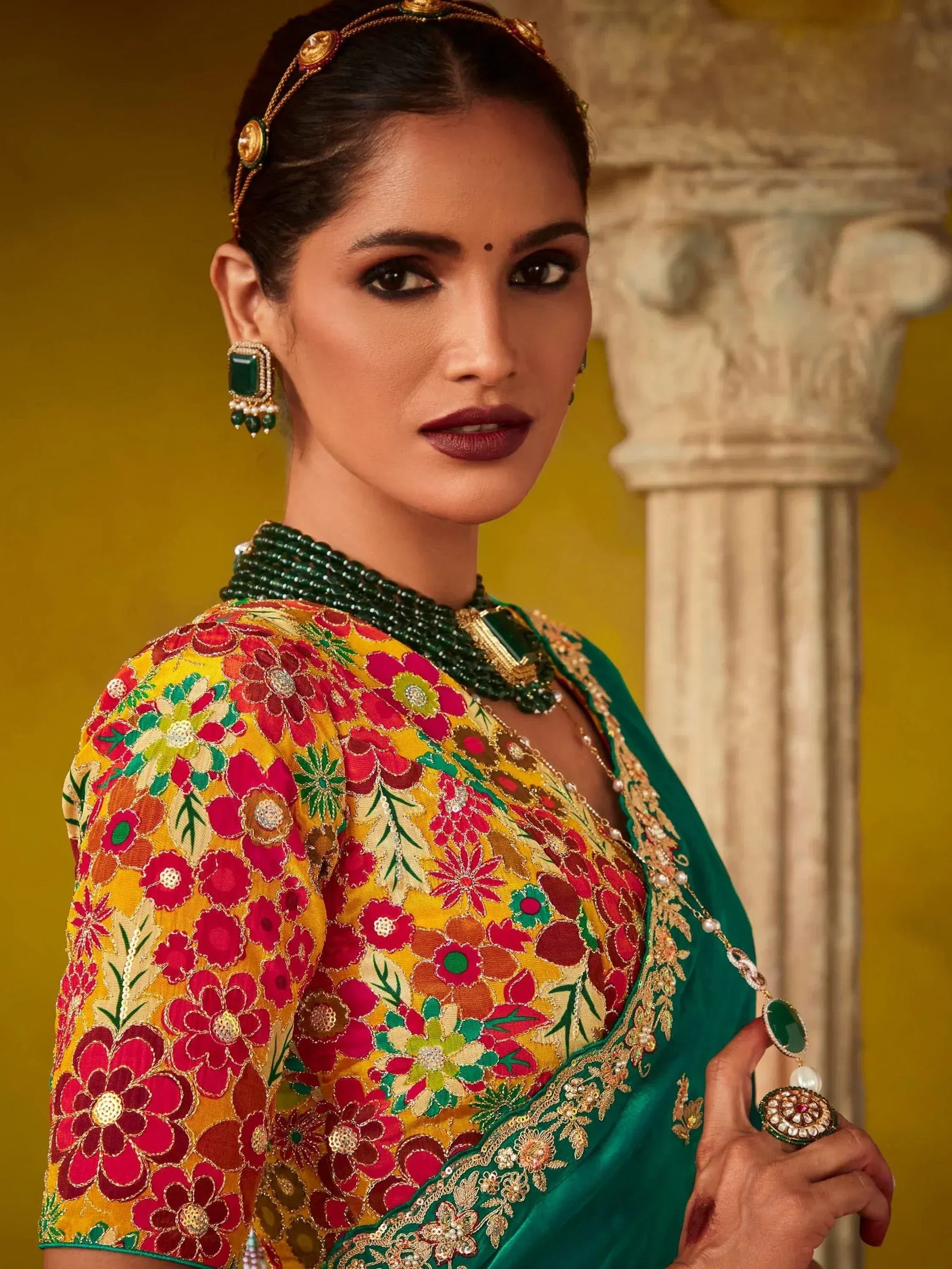 Woman in vibrant floral embroidered saree with green border, traditional Indian jewelry, and temple backdrop