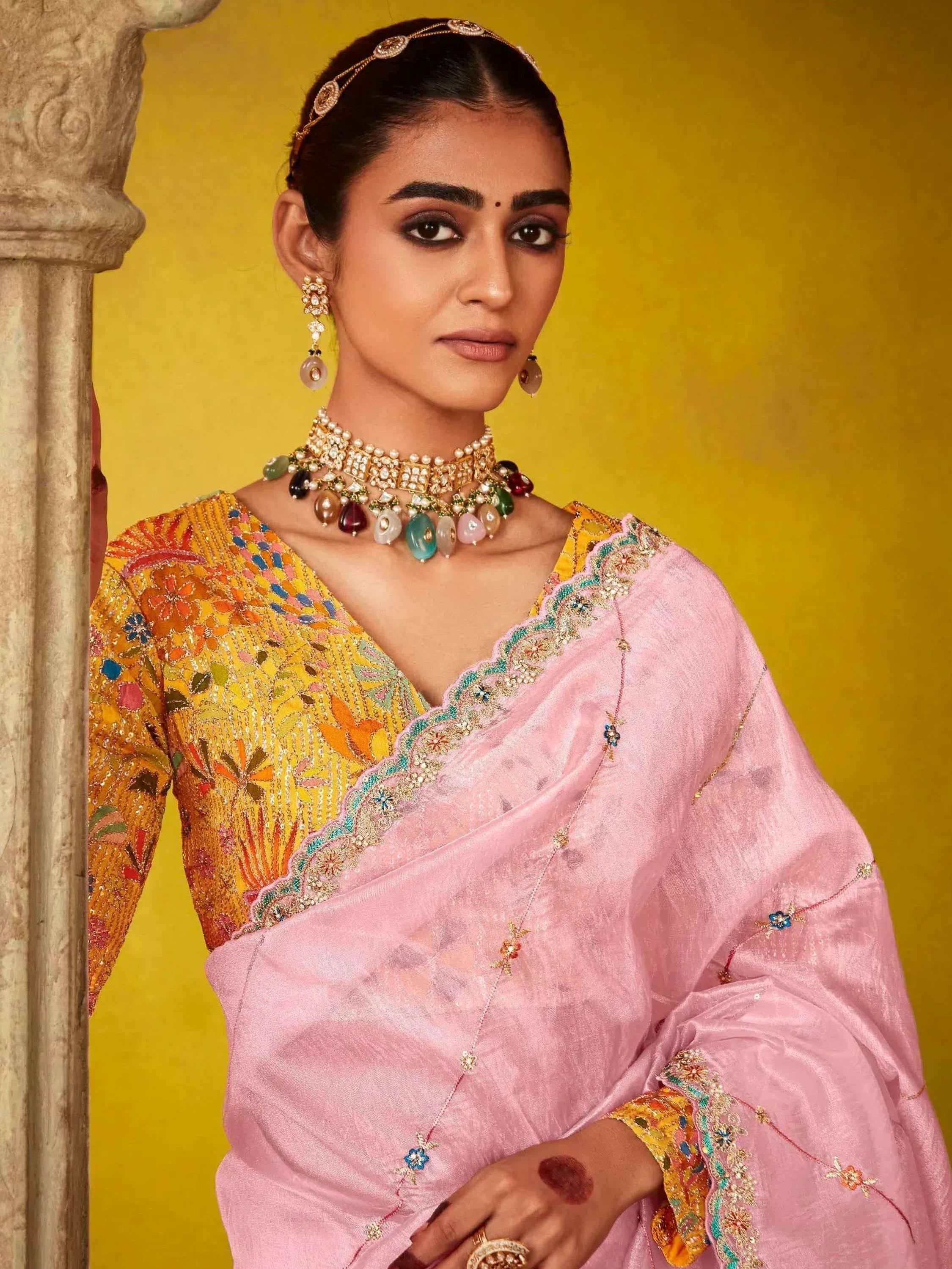 Woman in pink saree with floral embroidered blouse, traditional Indian jewelry, and henna, against yellow backdrop
