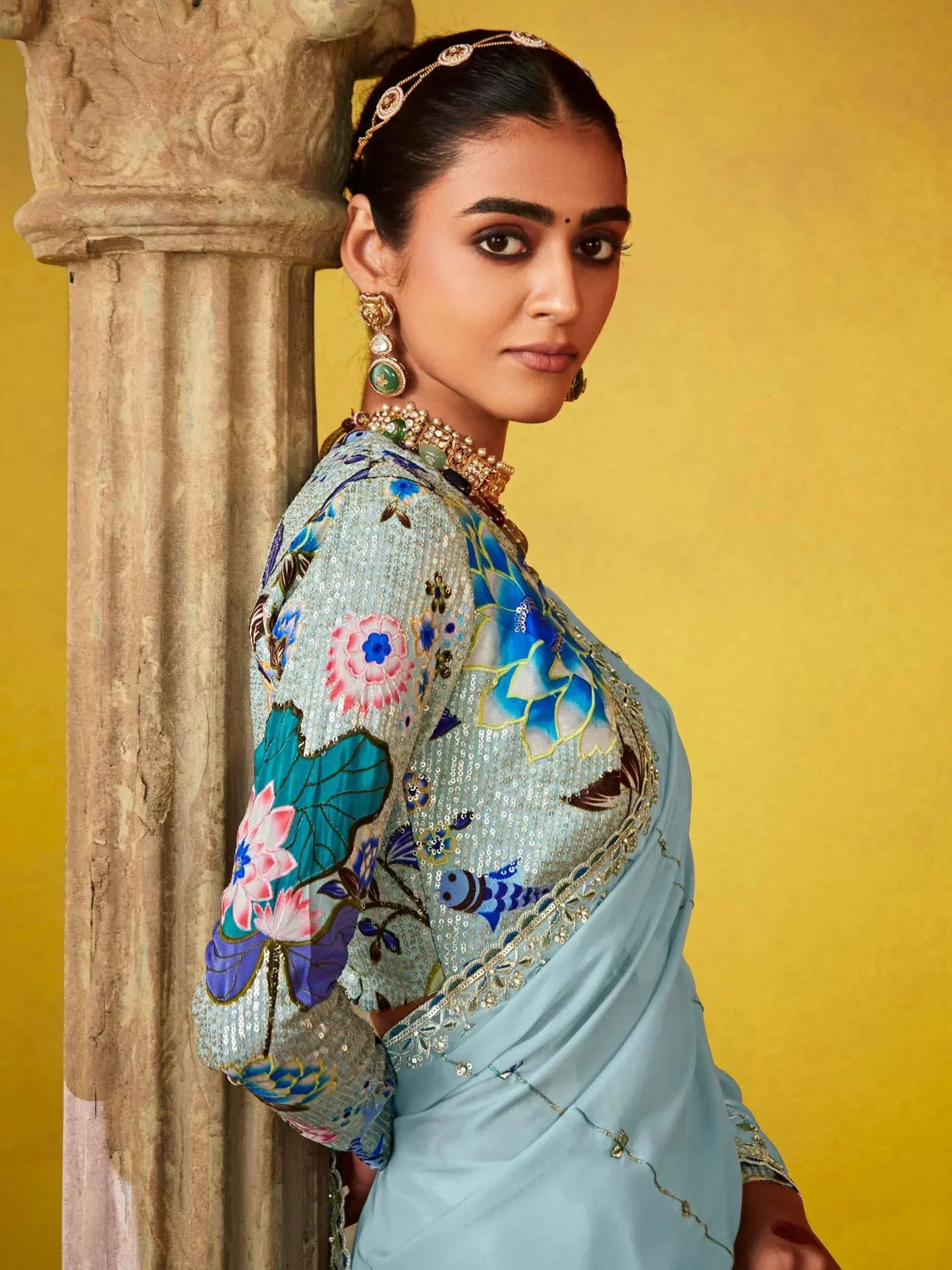 Woman in blue floral embroidered saree with gold jewelry, leaning against stone pillar