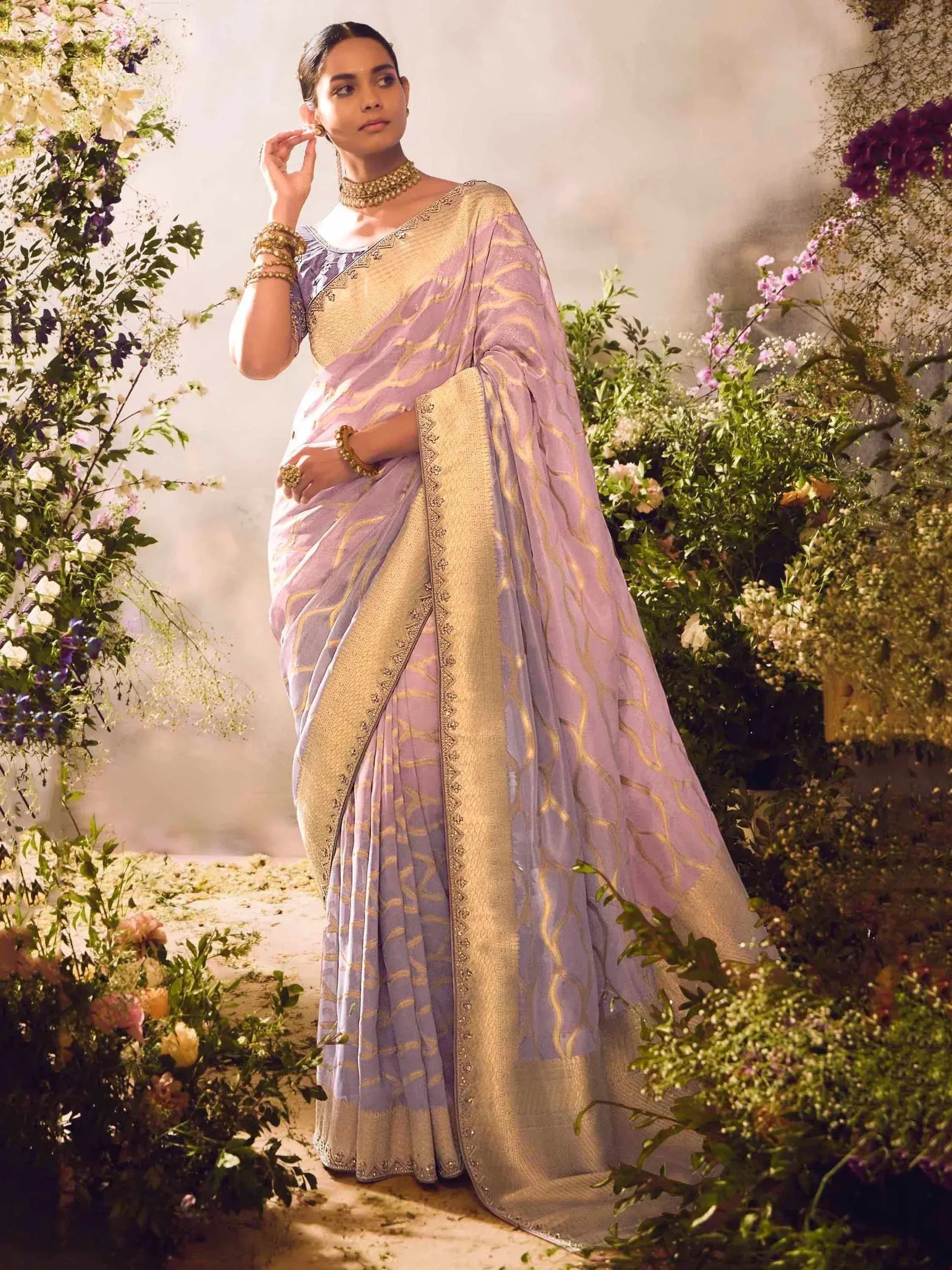 Lavender and gold designer saree with intricate embroidery, styled with traditional jewelry in a floral garden setting.