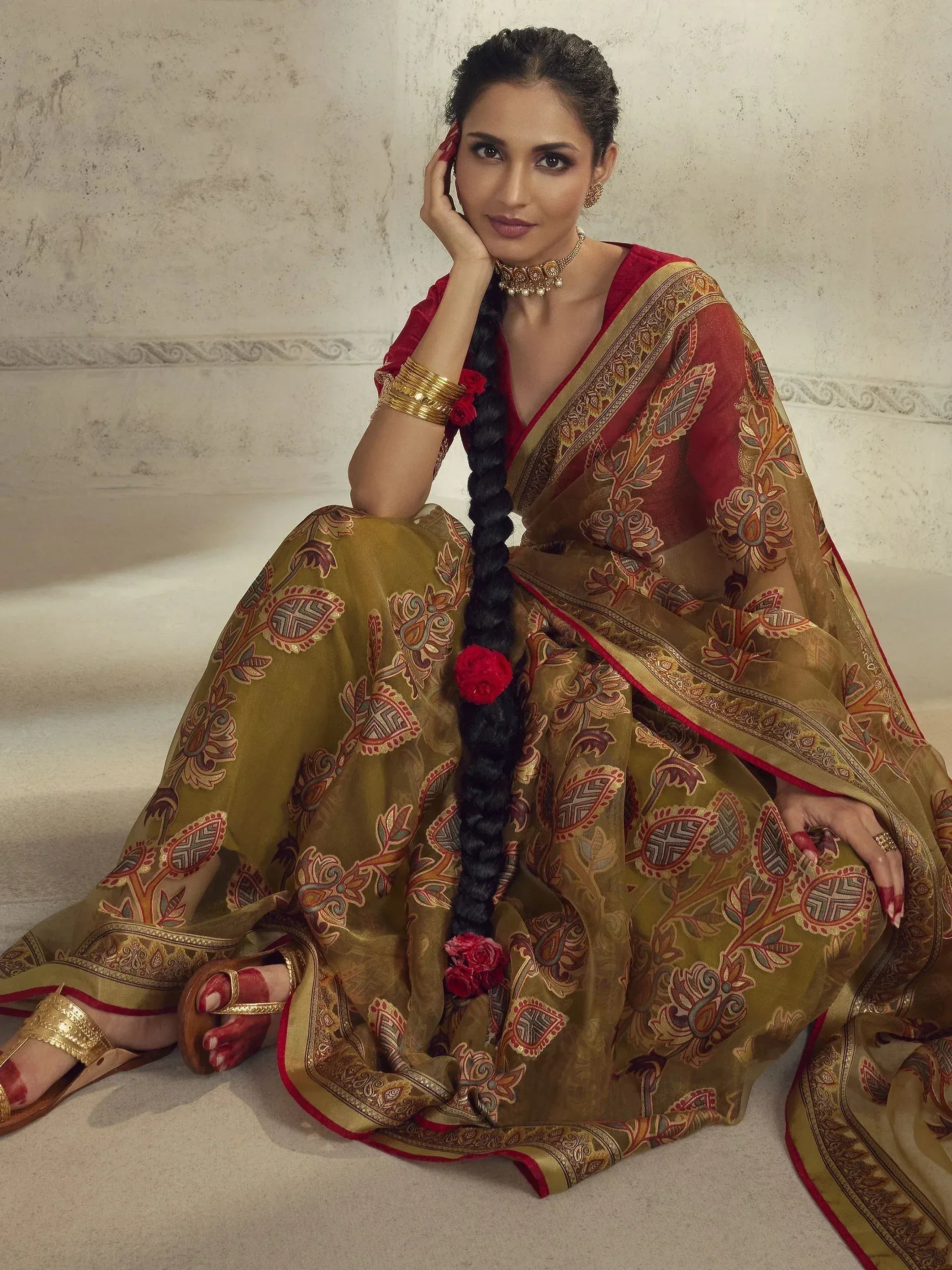 Woman in elegant red and gold printed saree with floral motifs, traditional jewelry, and braided hair adorned with roses.