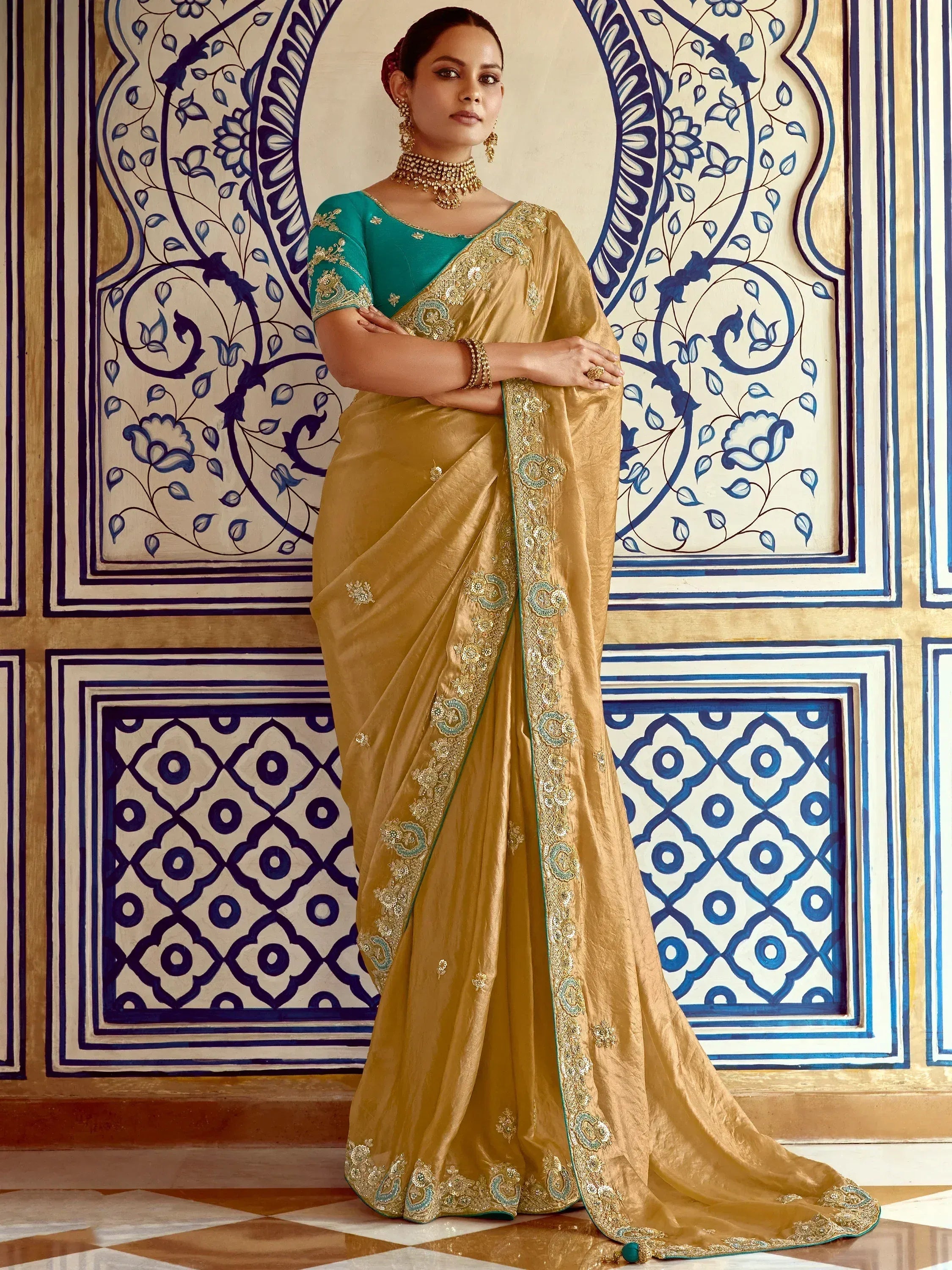 Golden saree with teal blouse, intricate embroidery, and traditional Indian jewelry against ornate blue patterns.
