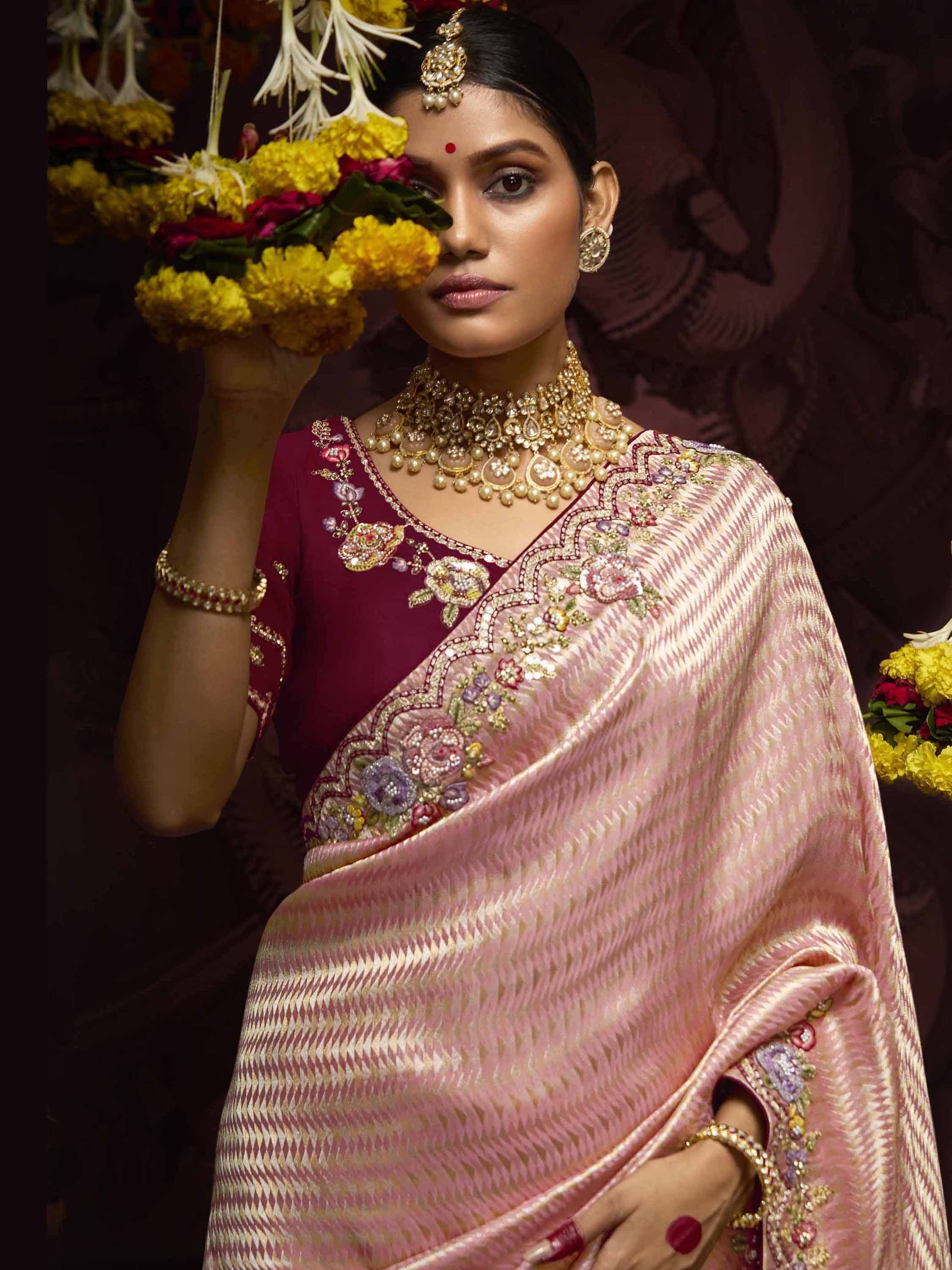 Elegant woman in pink embroidered saree with maroon blouse, gold jewelry, and floral garland, showcasing Indian ethnic wear.