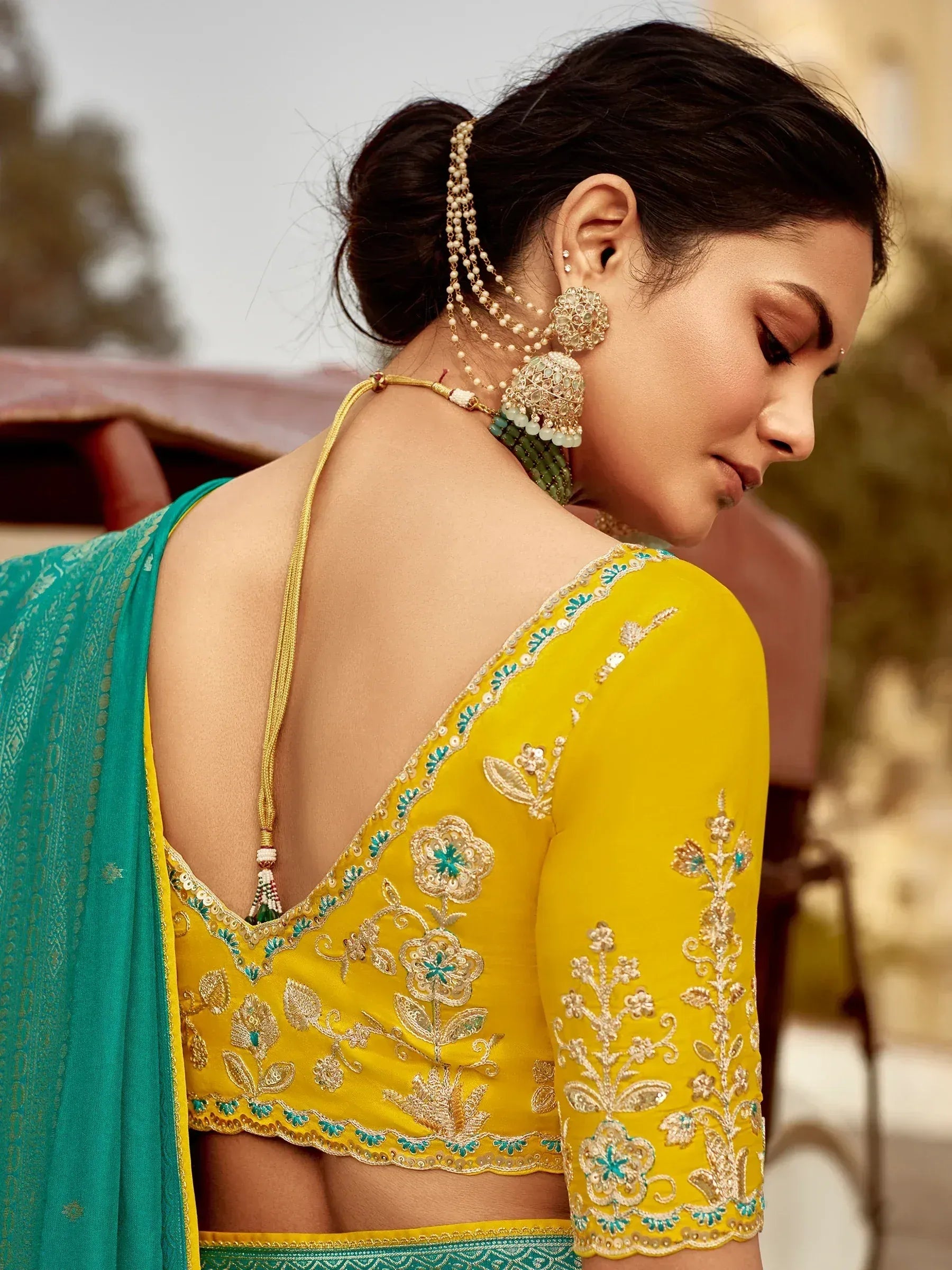 Yellow embroidered blouse with backless design, paired with teal saree, floral gold embellishments, and traditional jewelry.