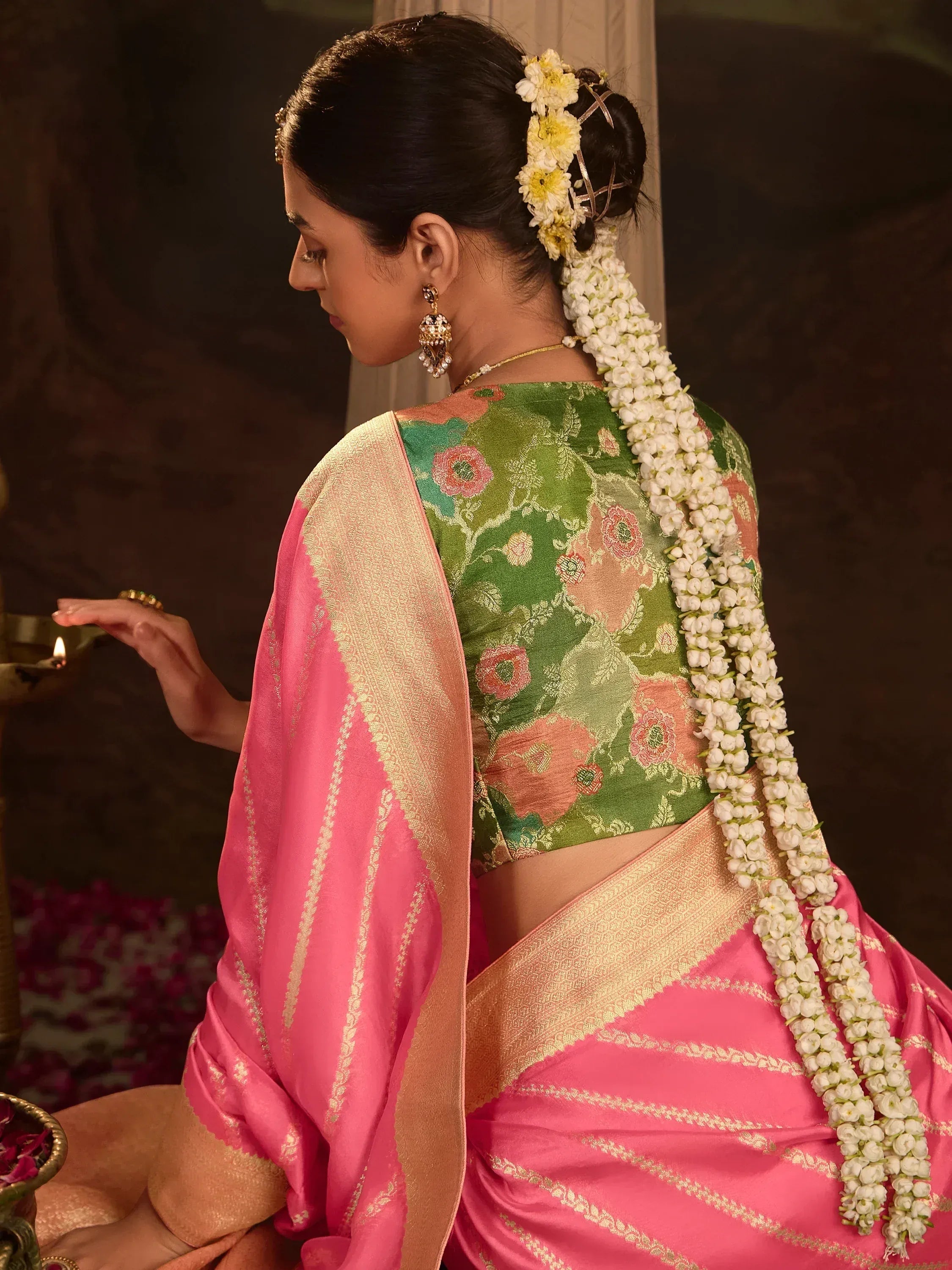 Woman in pink saree with green floral blouse, jasmine gajra, and gold jewelry, holding oil lamp