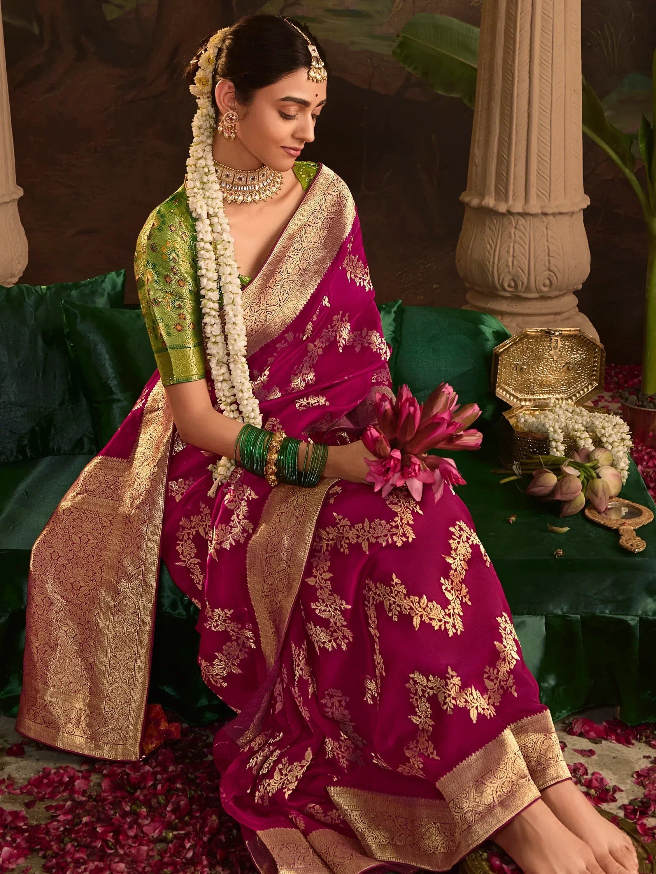 Elegant woman in magenta silk saree with gold embroidery, green blouse, floral garland, and traditional jewelry, seated on green velvet with rose petals.