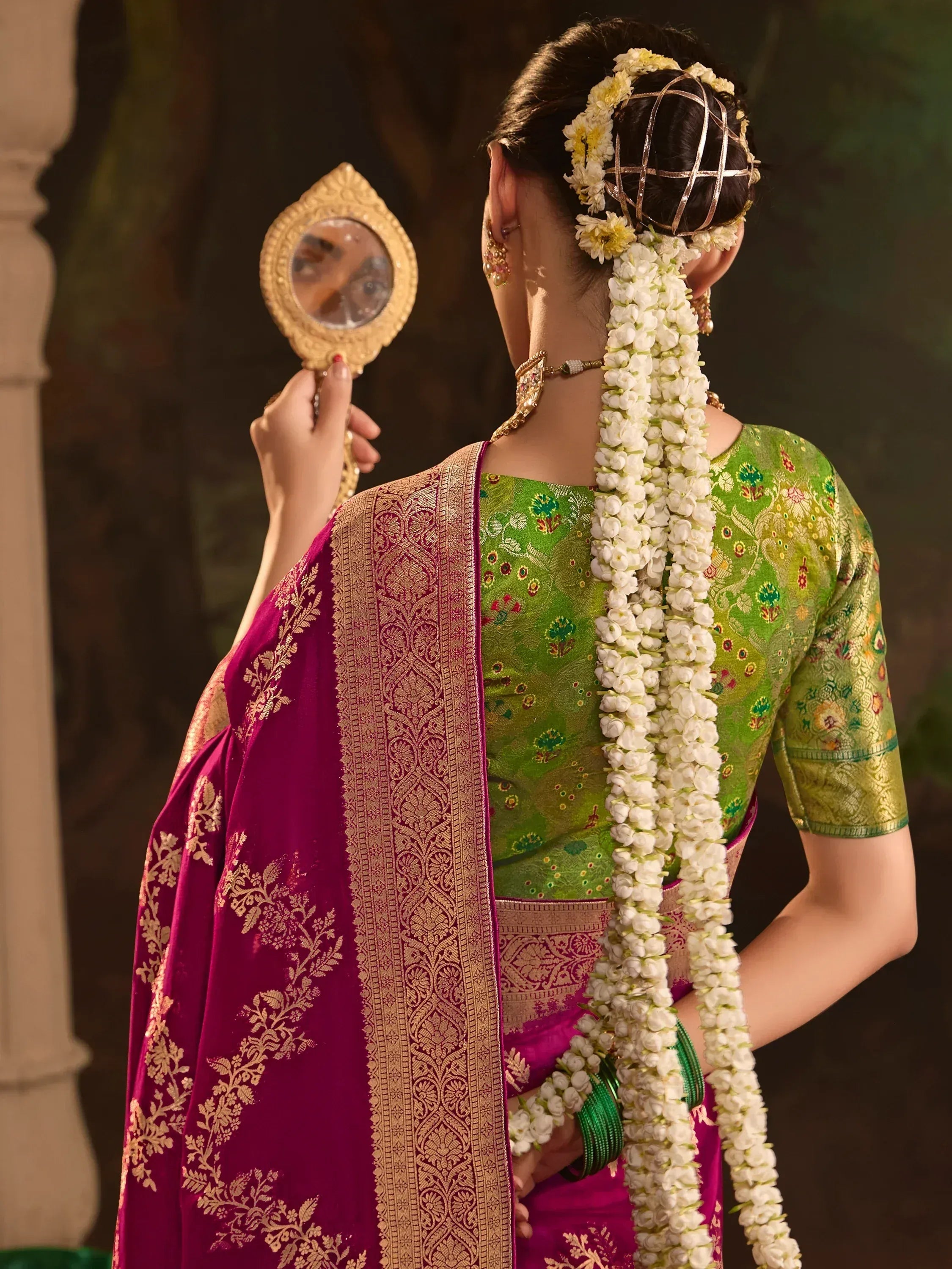 Woman in green and magenta saree with floral hair adornment holding ornate mirror