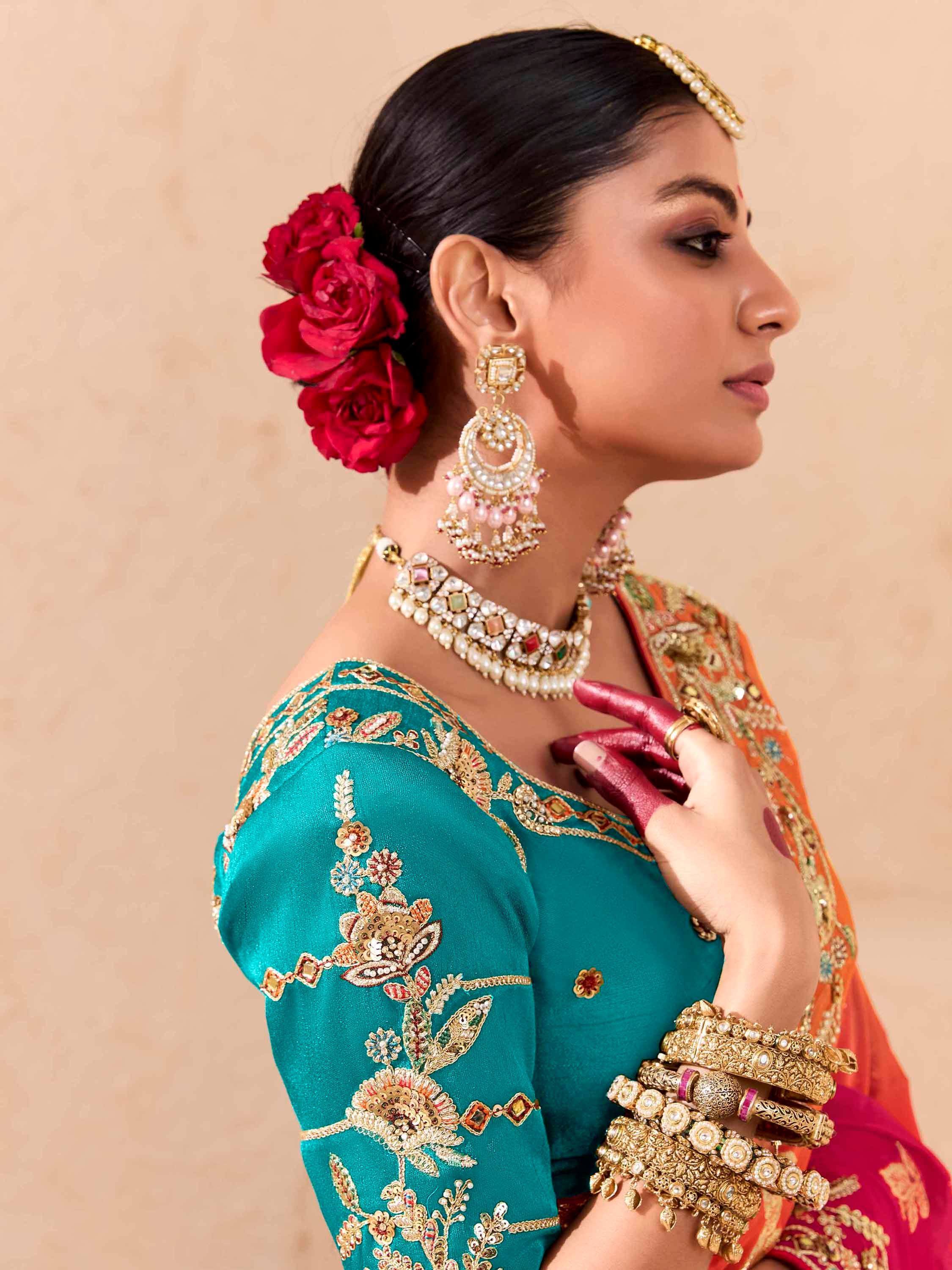 Woman in teal embroidered blouse with red roses, gold jewelry, and orange saree for Indian ethnic wear