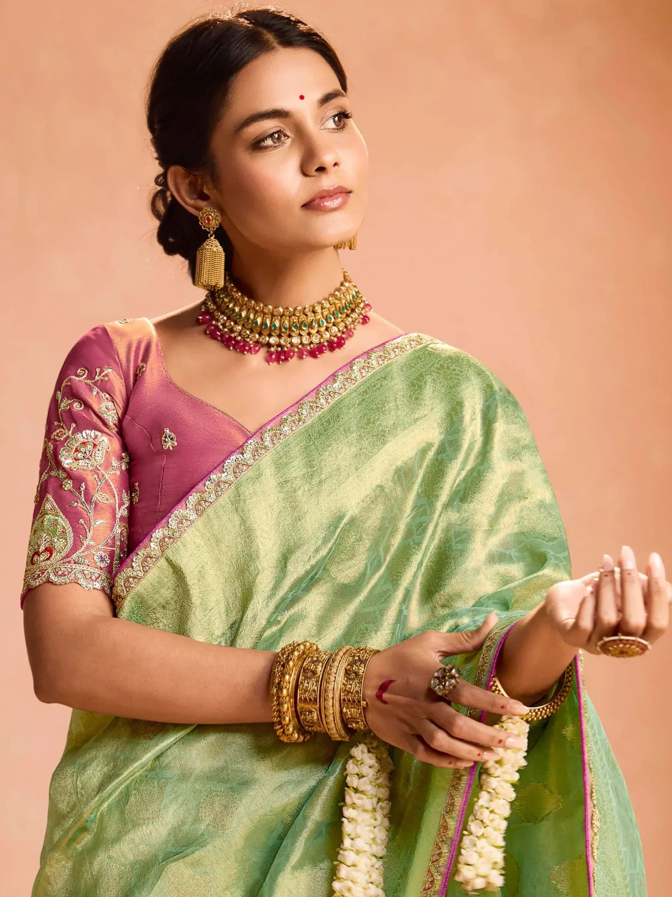 Elegant woman in green silk saree with pink embroidered blouse, traditional gold jewelry, and floral garland