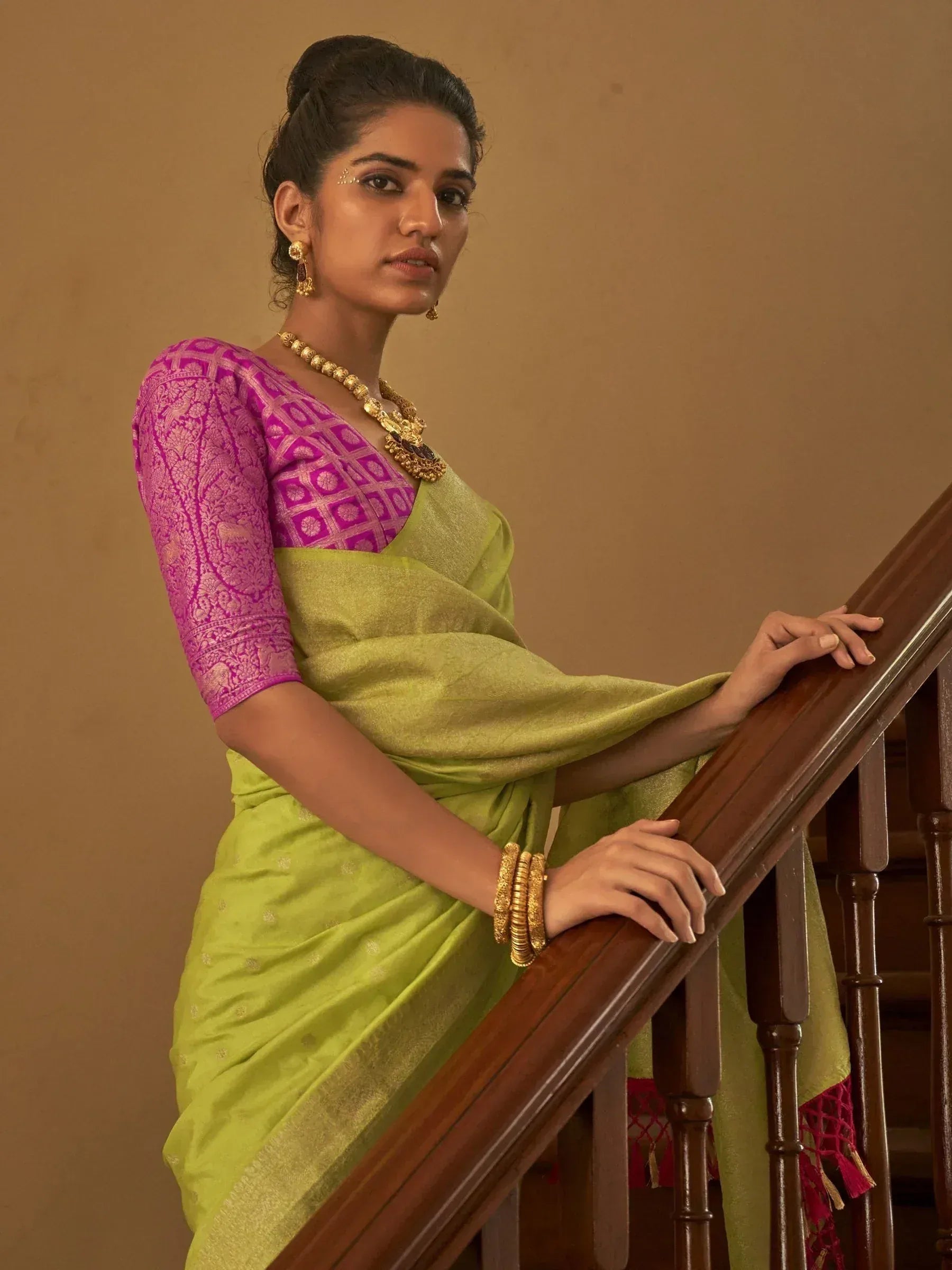 Woman in vibrant green saree with pink brocade blouse, gold jewelry, leaning on wooden staircase