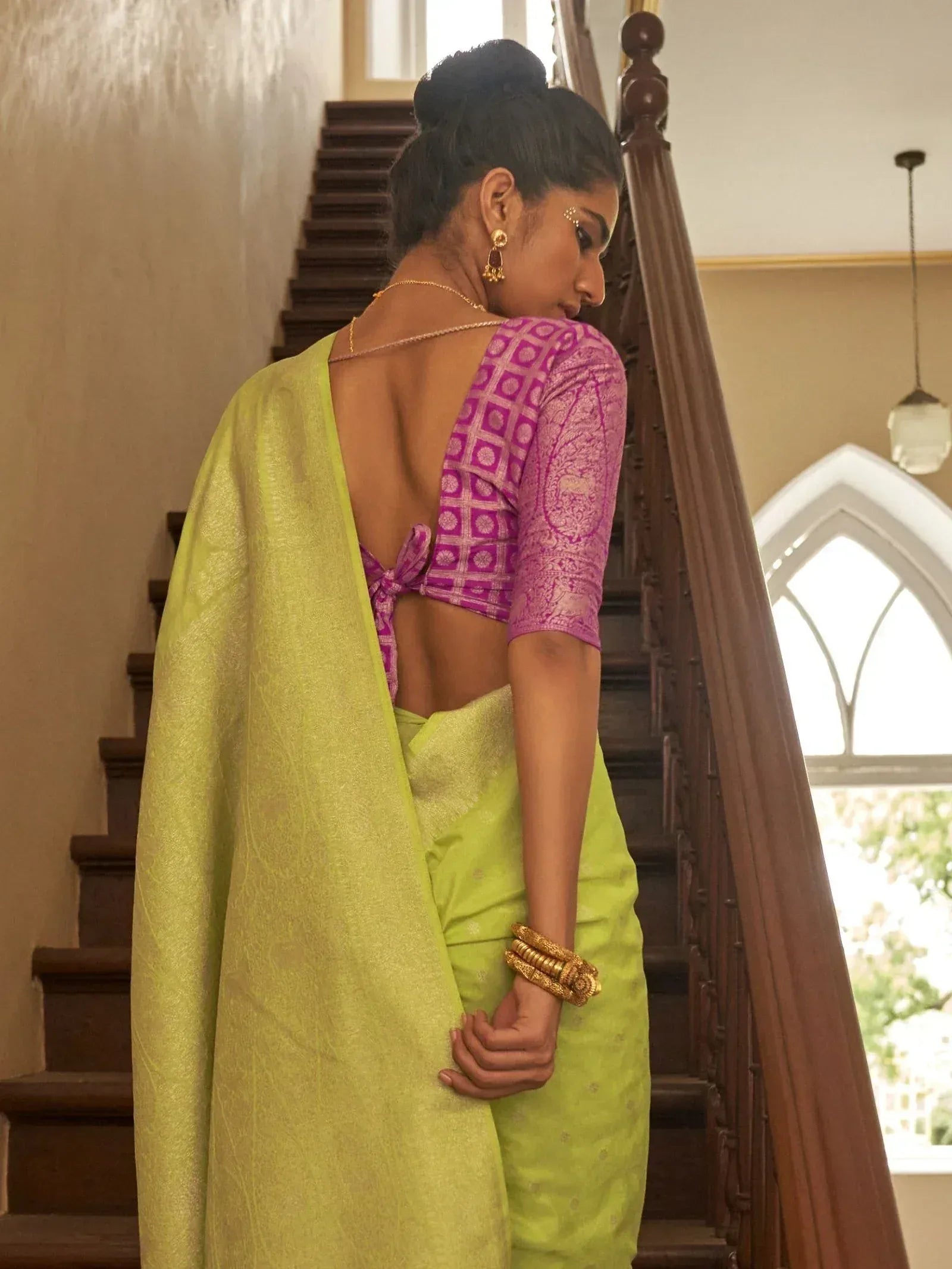 Woman in vibrant green saree with purple patterned blouse, standing by wooden staircase