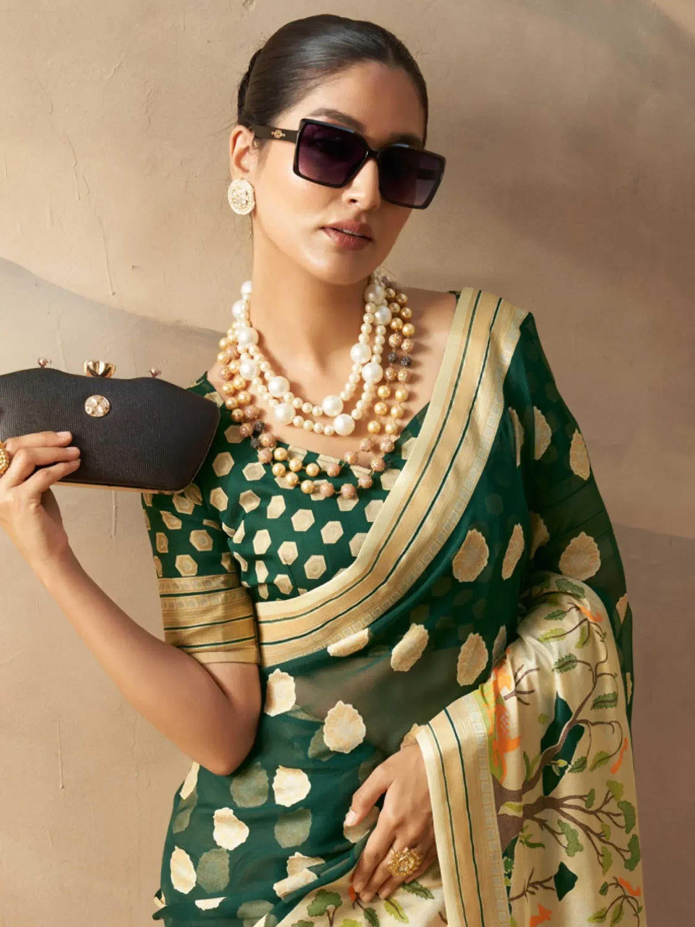 Woman in green floral saree with gold border, pearl necklace, and sunglasses, holding black clutch