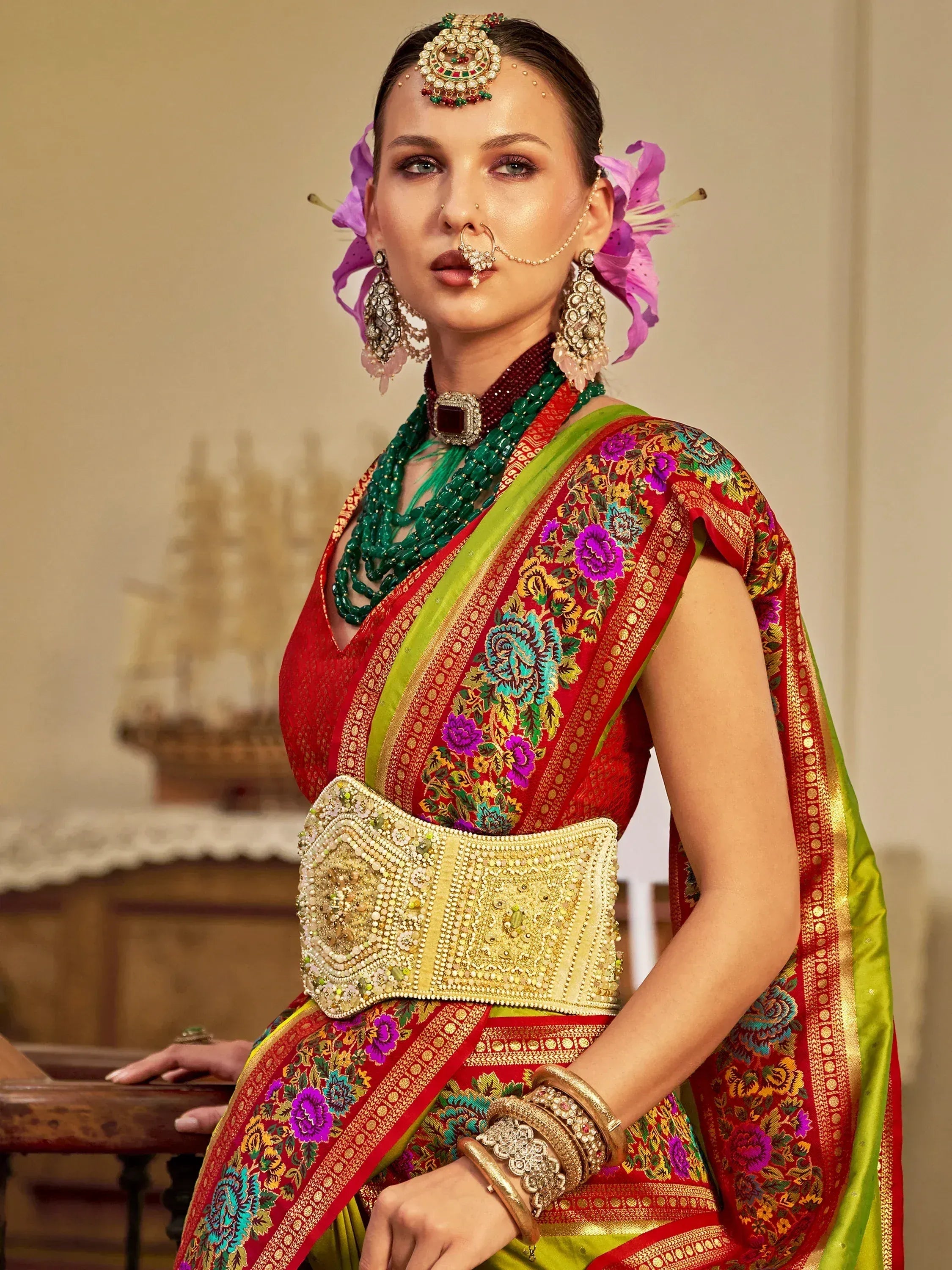 Woman in vibrant red and green embroidered saree with floral patterns, traditional Indian jewelry, and ornate waist belt