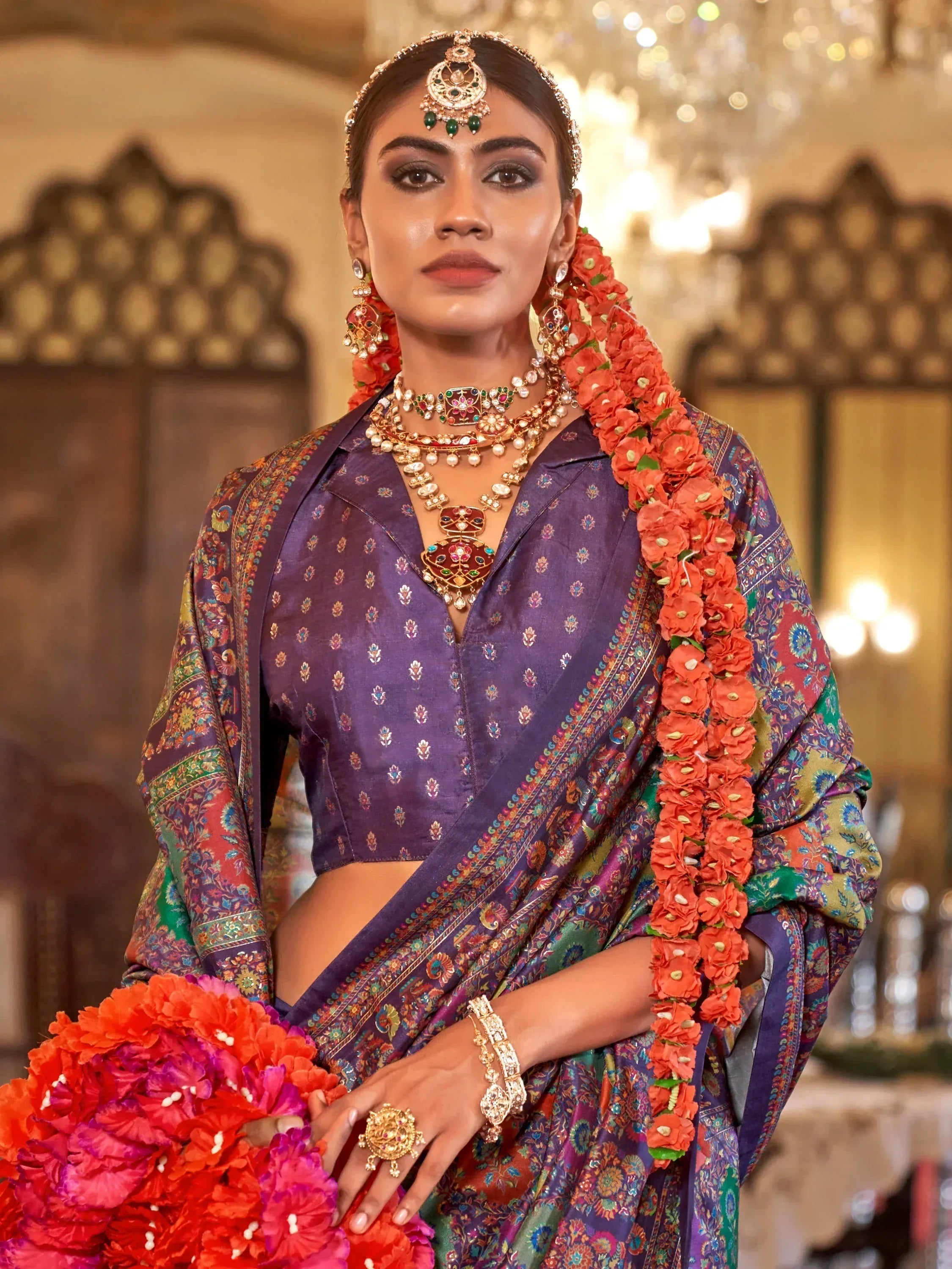 Elegant woman in purple ethnic saree with floral patterns, traditional jewelry, and vibrant orange garlands.
