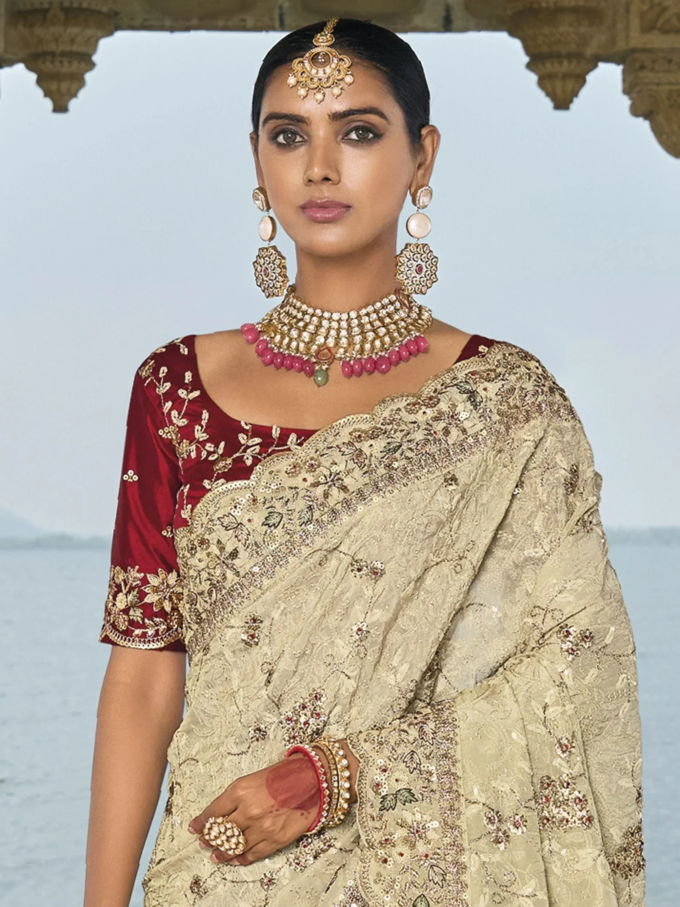 Elegant woman in beige embroidered saree with maroon blouse, traditional Indian jewelry, and temple backdrop