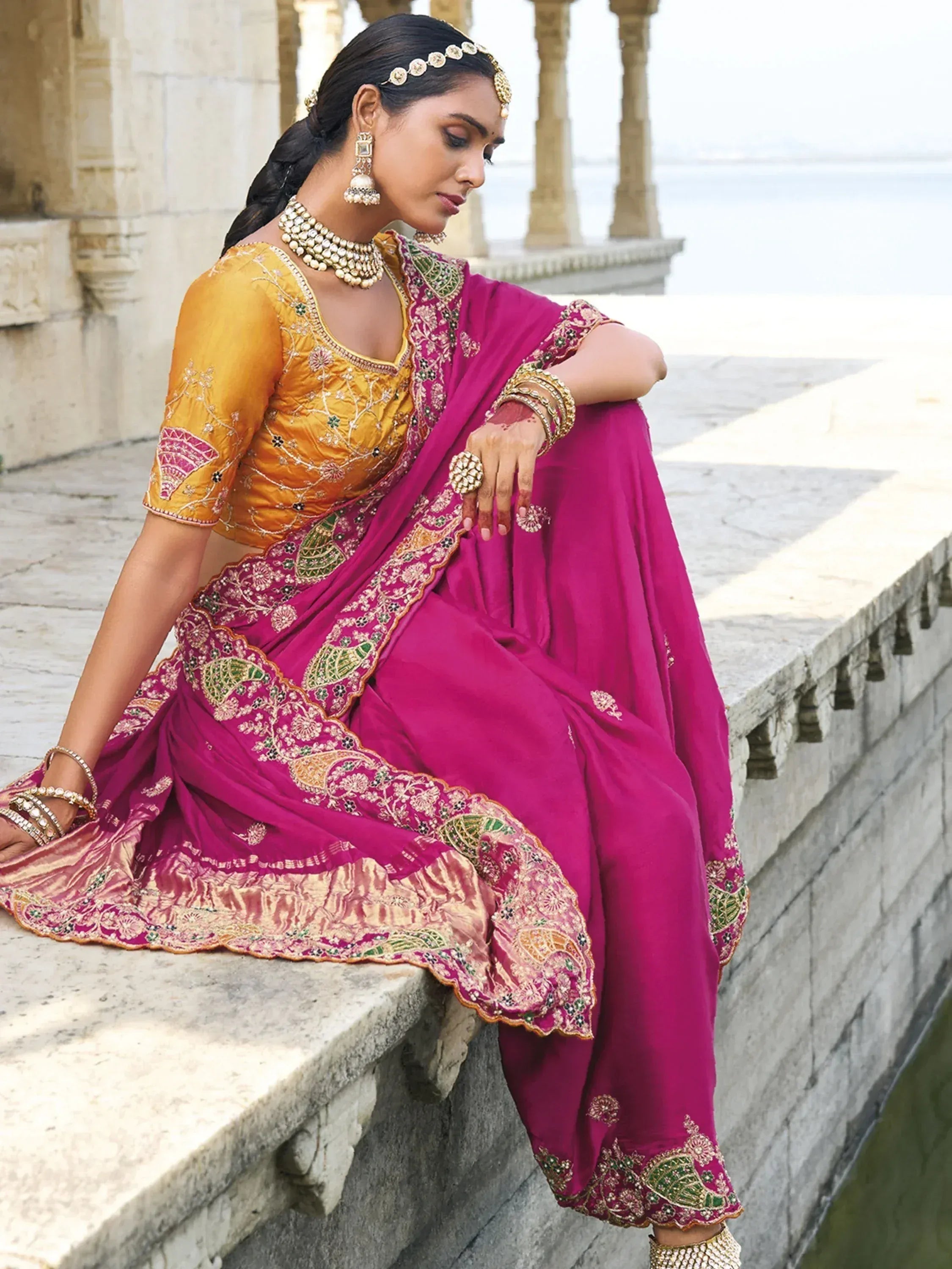 Woman in vibrant pink and gold embroidered Indian ethnic wear, sitting by a stone balcony with traditional jewelry.