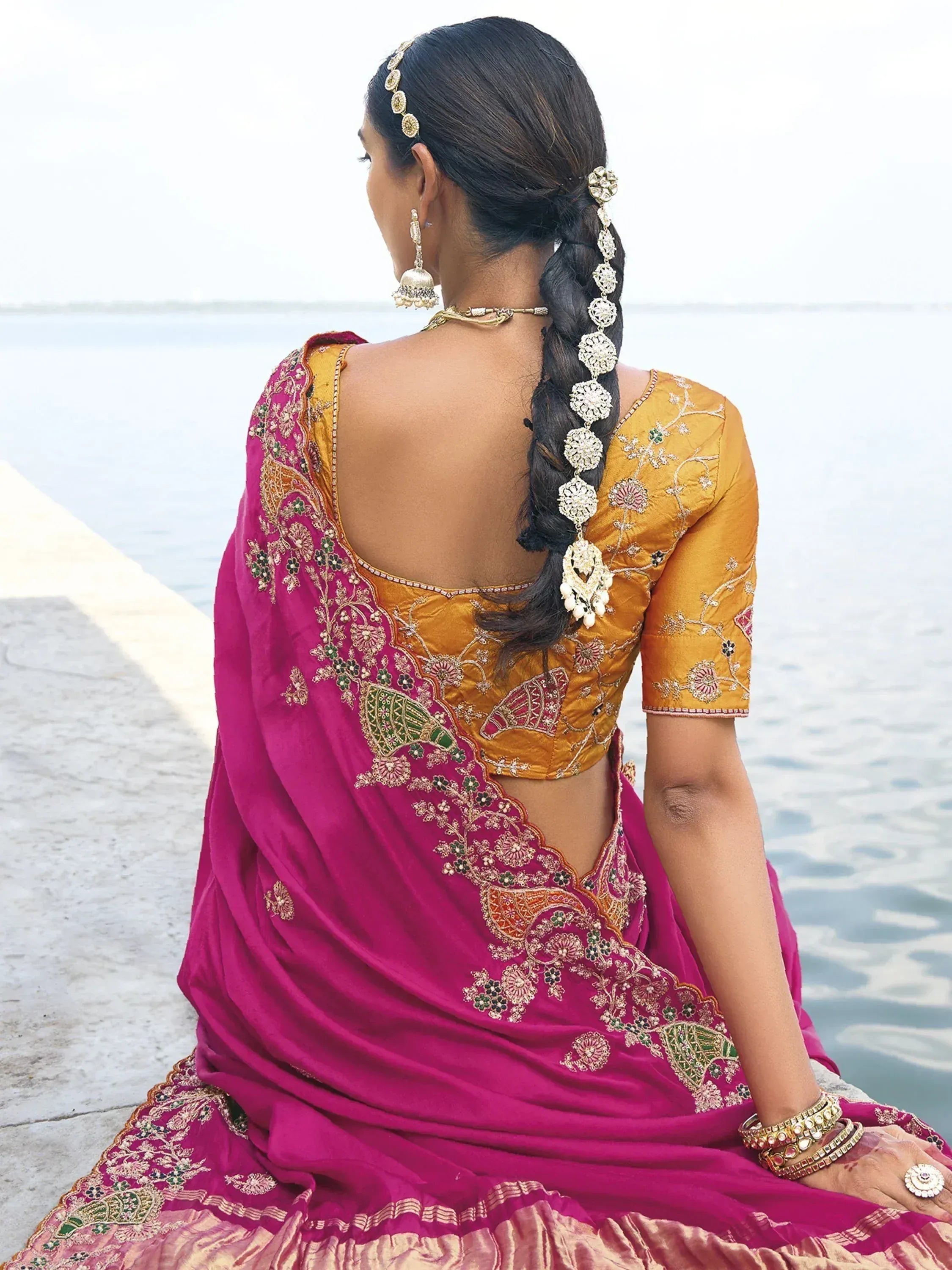 Woman in pink and gold embroidered saree with backless blouse, sitting by water, adorned with traditional jewelry