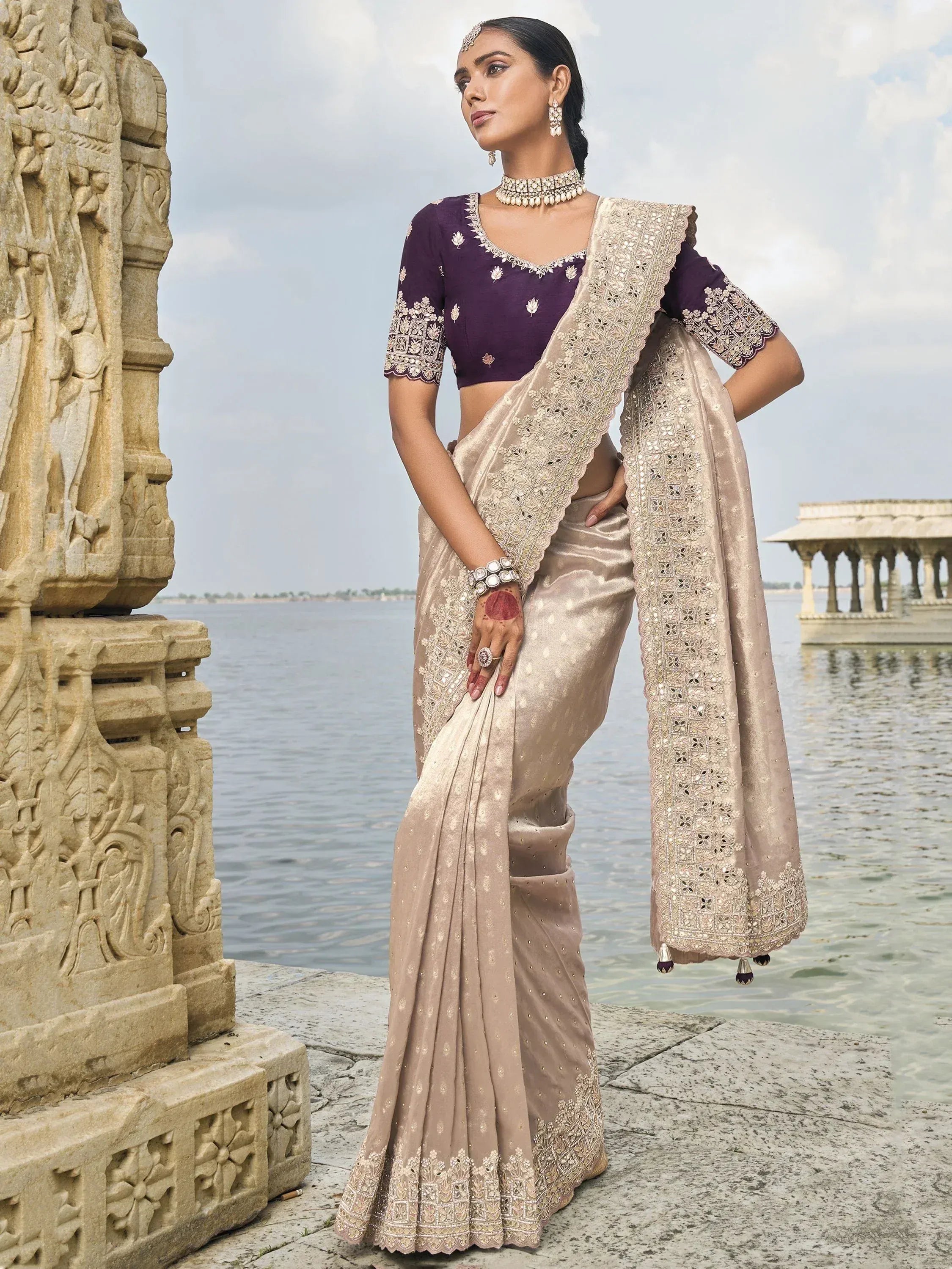 Elegant beige saree with gold embroidery and purple blouse, styled with traditional jewelry near water