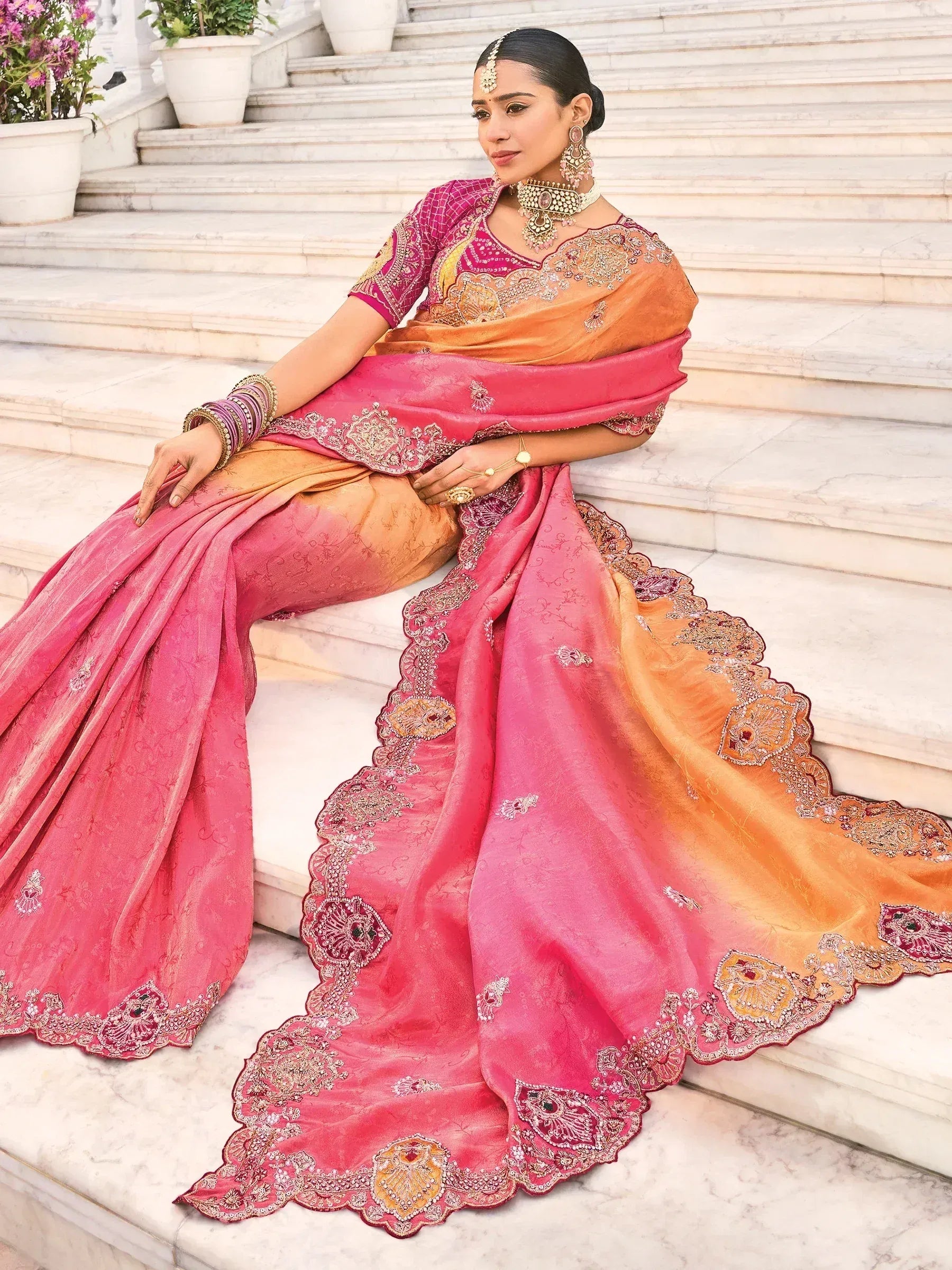 Pink and orange embroidered saree with gold detailing, worn by woman on marble steps