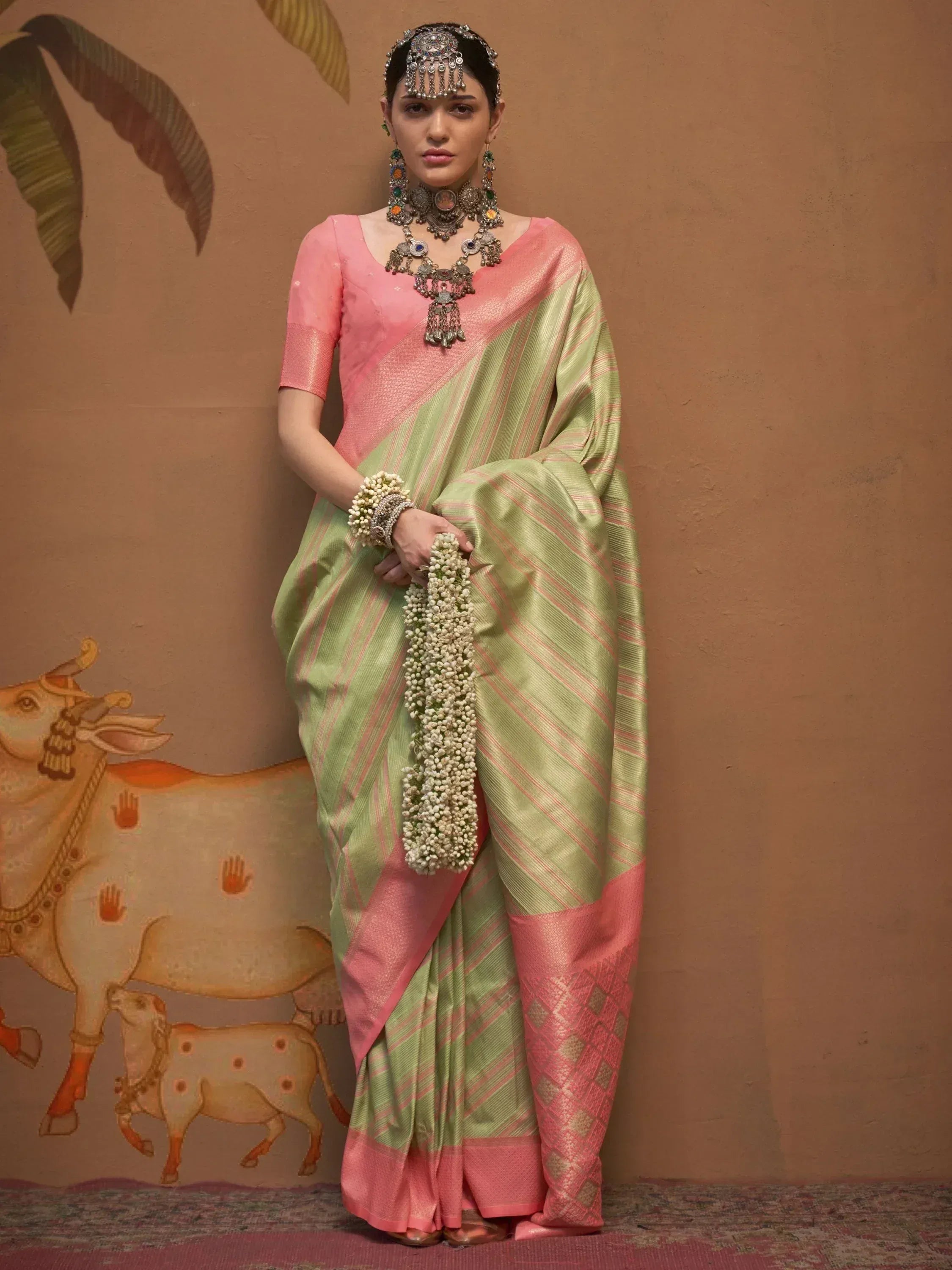 Woman in green and pink striped saree with traditional jewelry, holding jasmine garland, against painted wall
