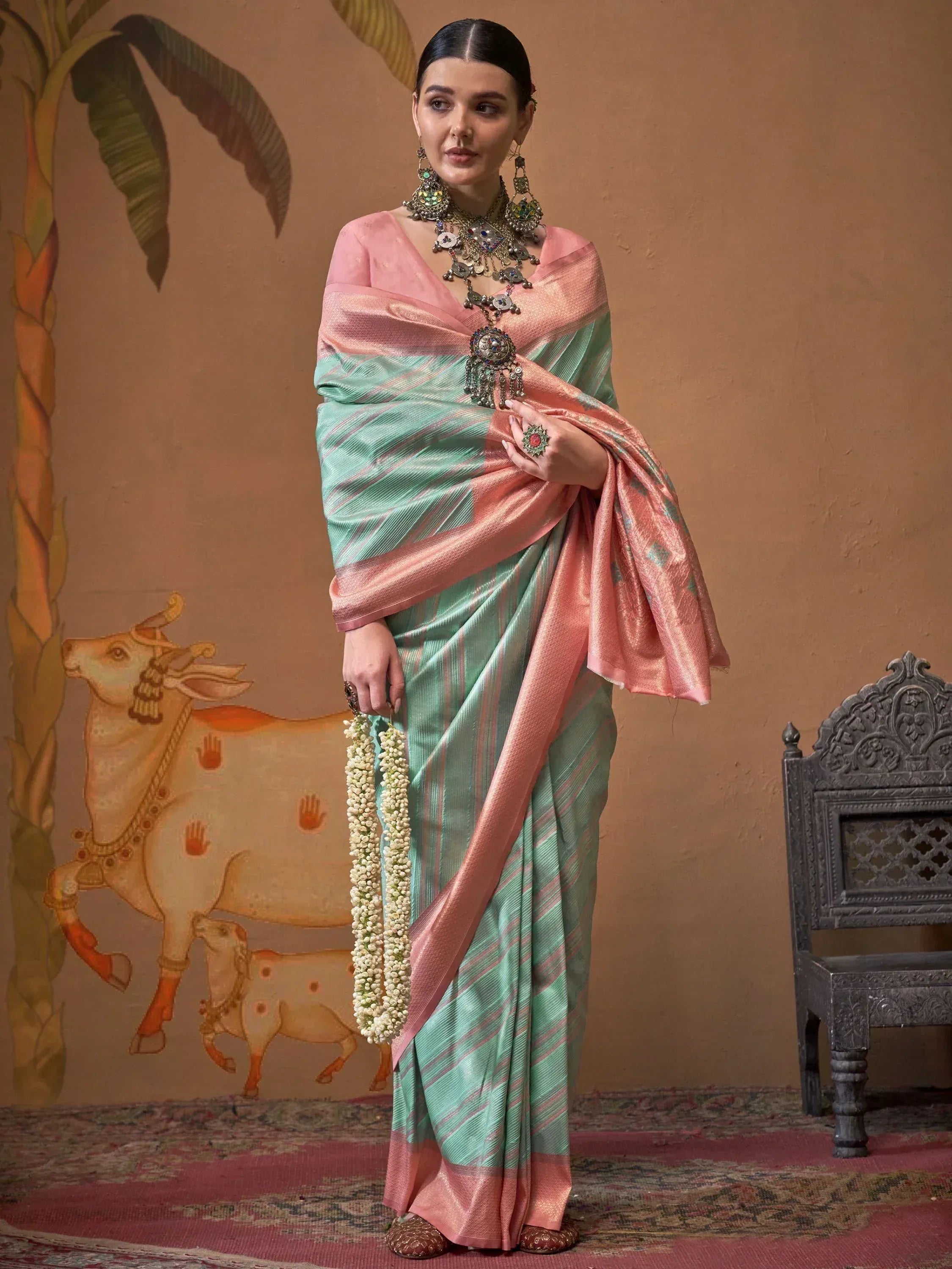 Woman in mint green and pink striped saree with traditional jewelry, holding floral garland, ethnic wear