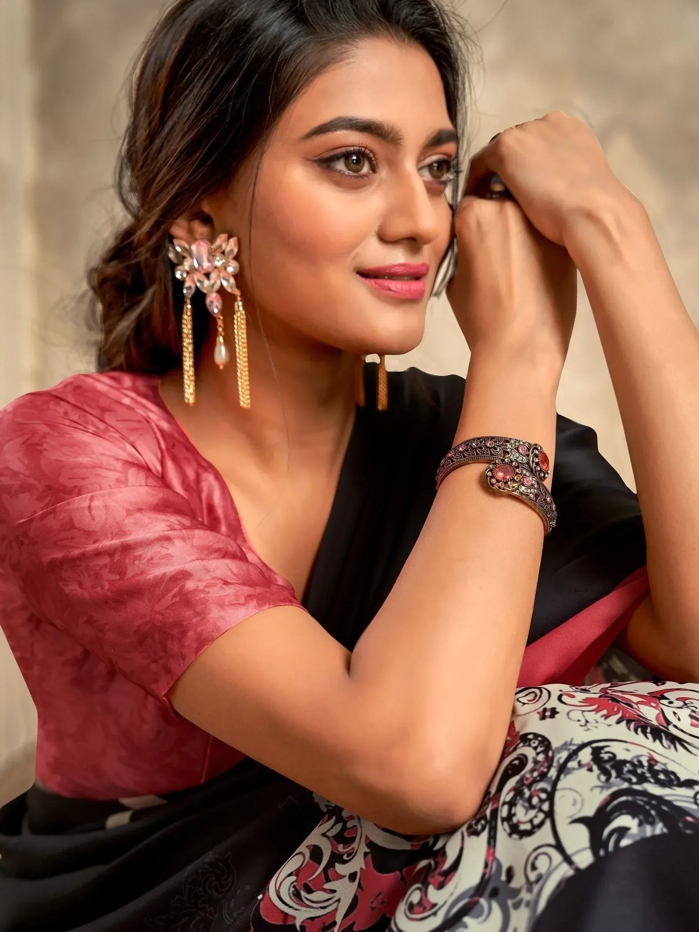 Woman in pink blouse and black saree with floral print, wearing ornate pink and gold earrings and bracelet