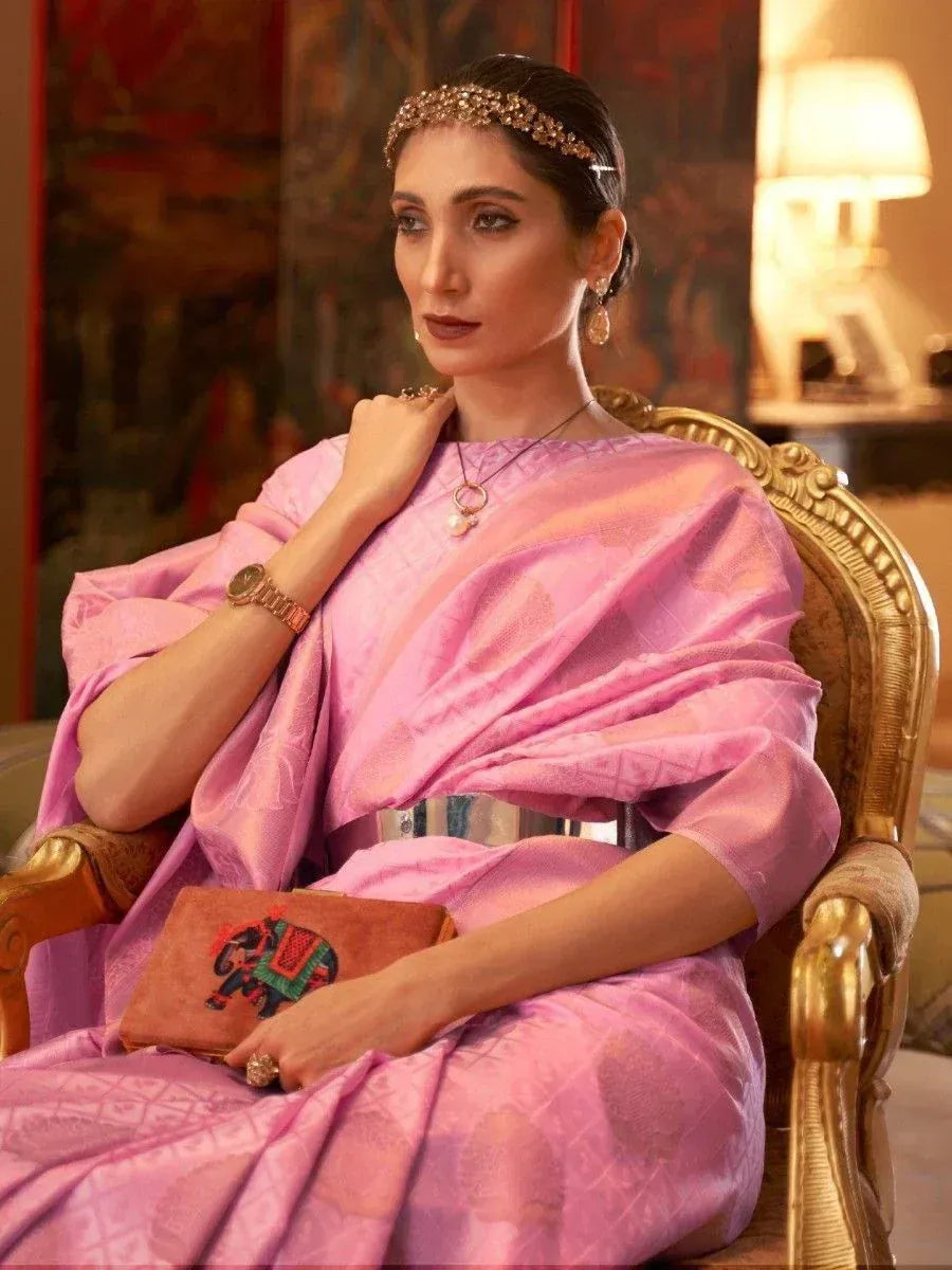 Woman in elegant pink ethnic gown with embroidery, gold accessories, and elephant clutch, seated in ornate chair
