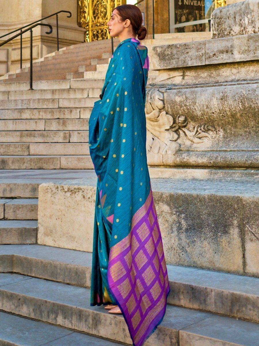 Woman in teal and purple ethnic saree with gold dots, standing on stone steps