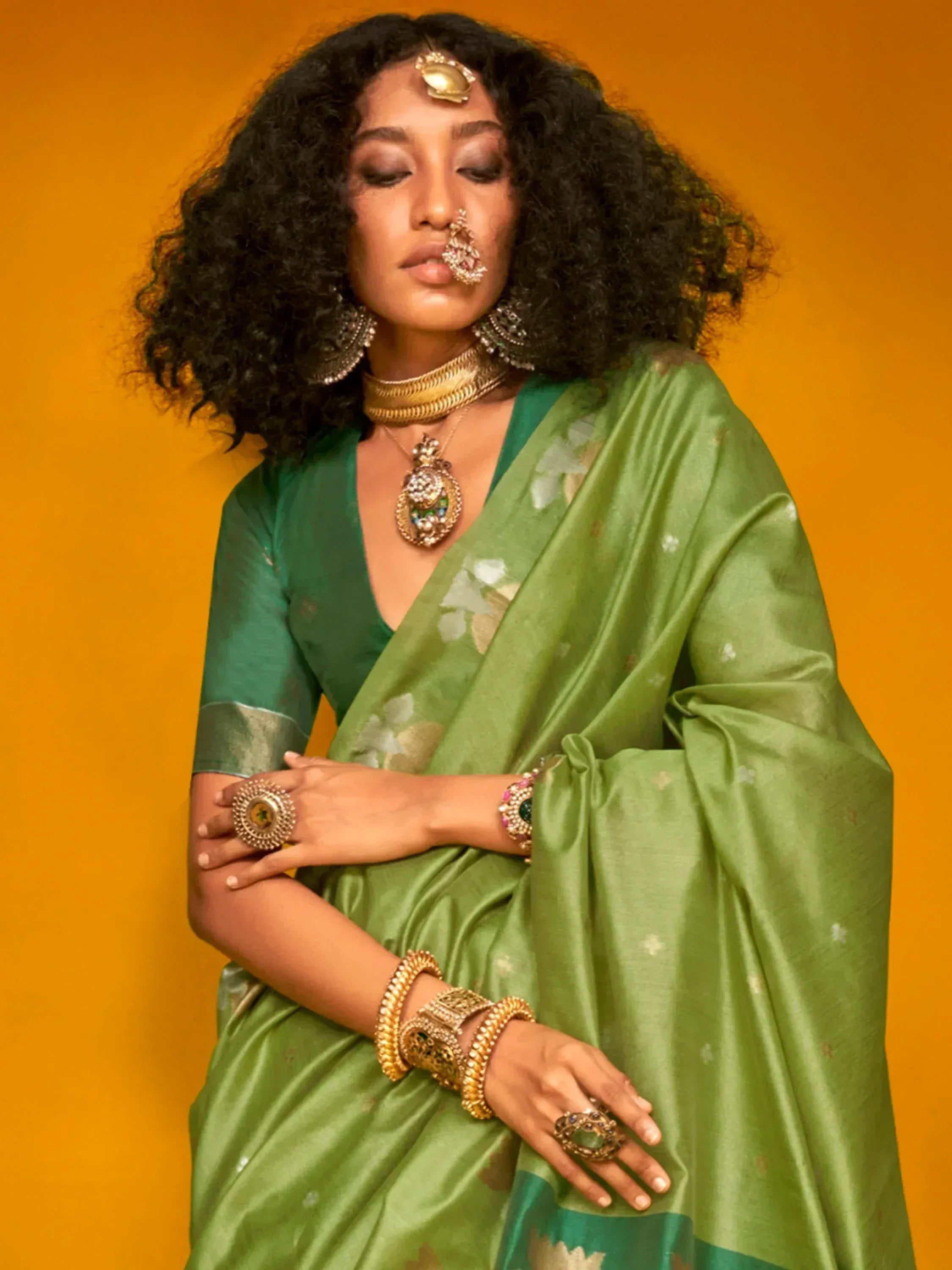 Woman in green silk saree with floral print, traditional Indian jewelry, and bold makeup against orange background