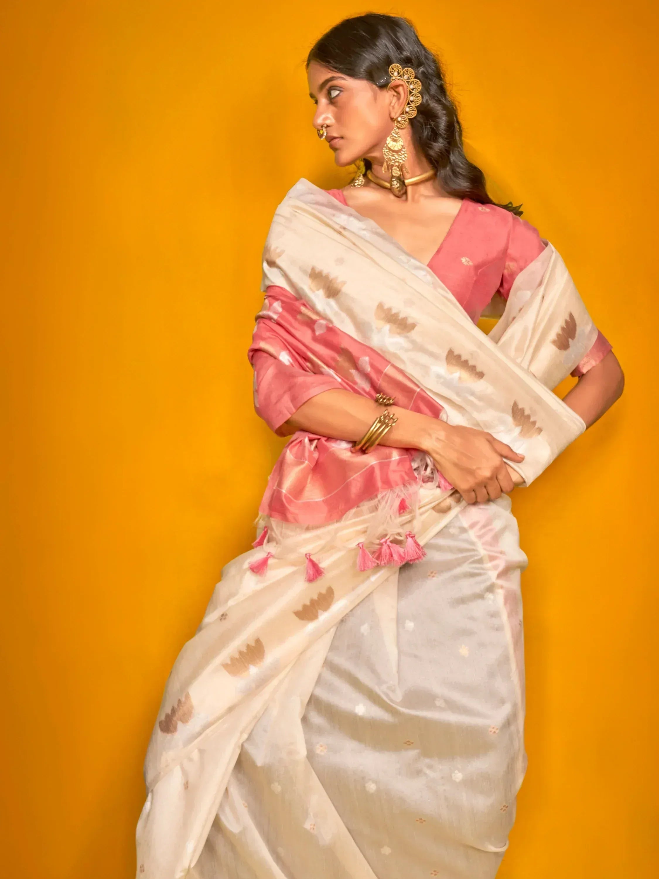 Woman in elegant white saree with pink blouse and floral print, styled with gold jewelry against yellow backdrop