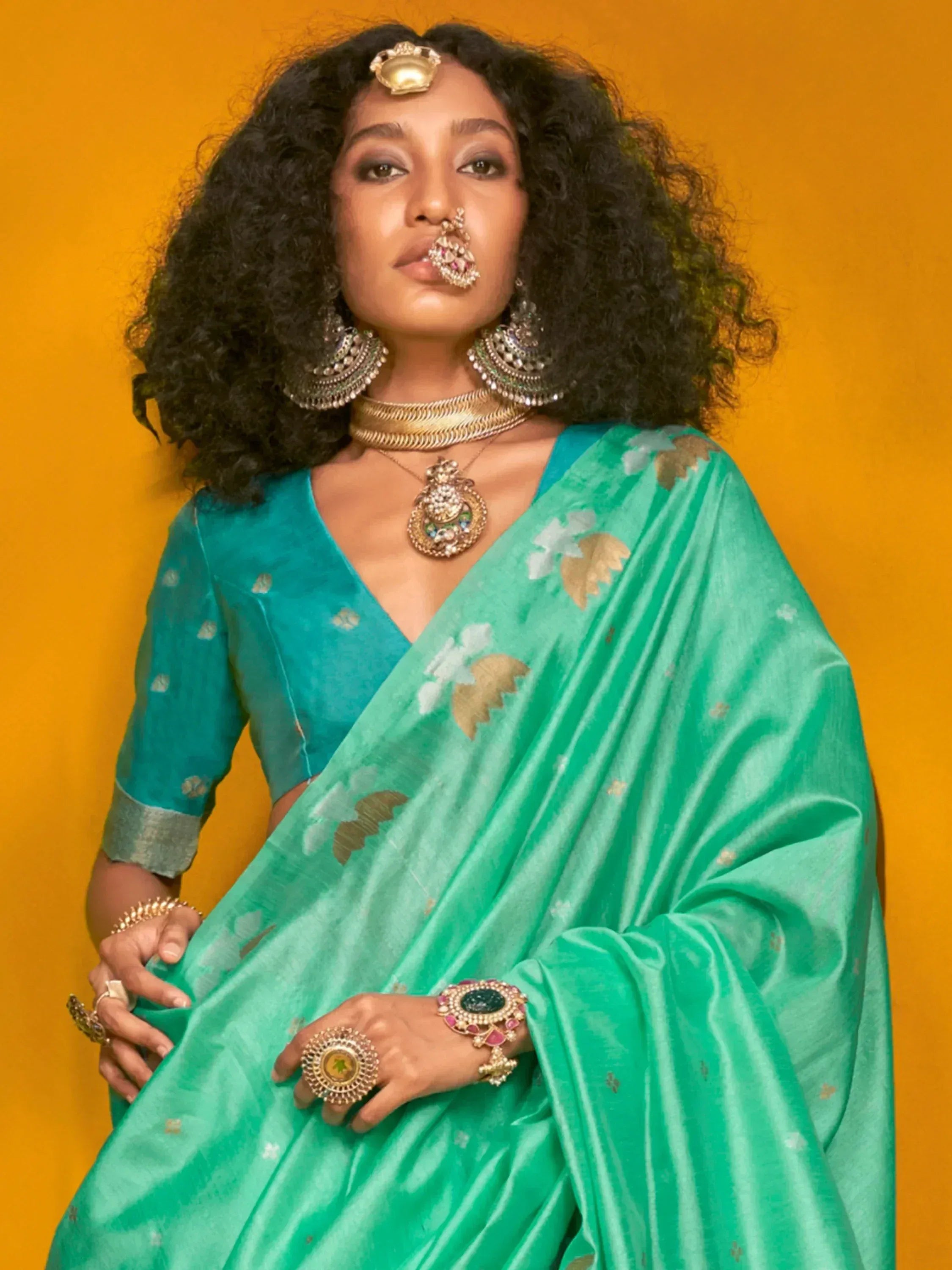 Woman in turquoise saree with gold floral motifs, traditional Indian jewelry, against orange backdrop