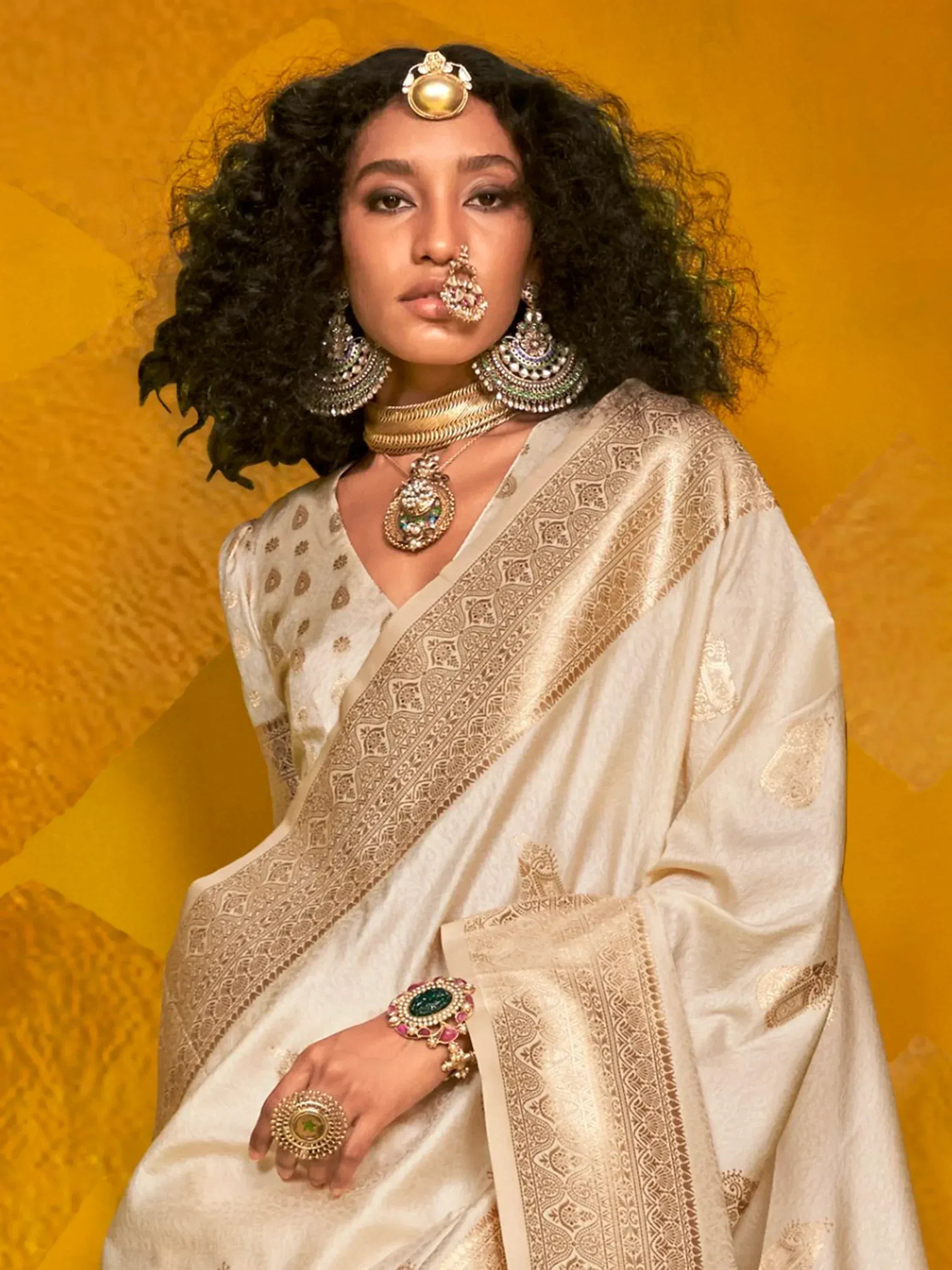 Elegant woman in cream saree with gold embroidery, traditional Indian jewelry, and bold curly hair against golden backdrop
