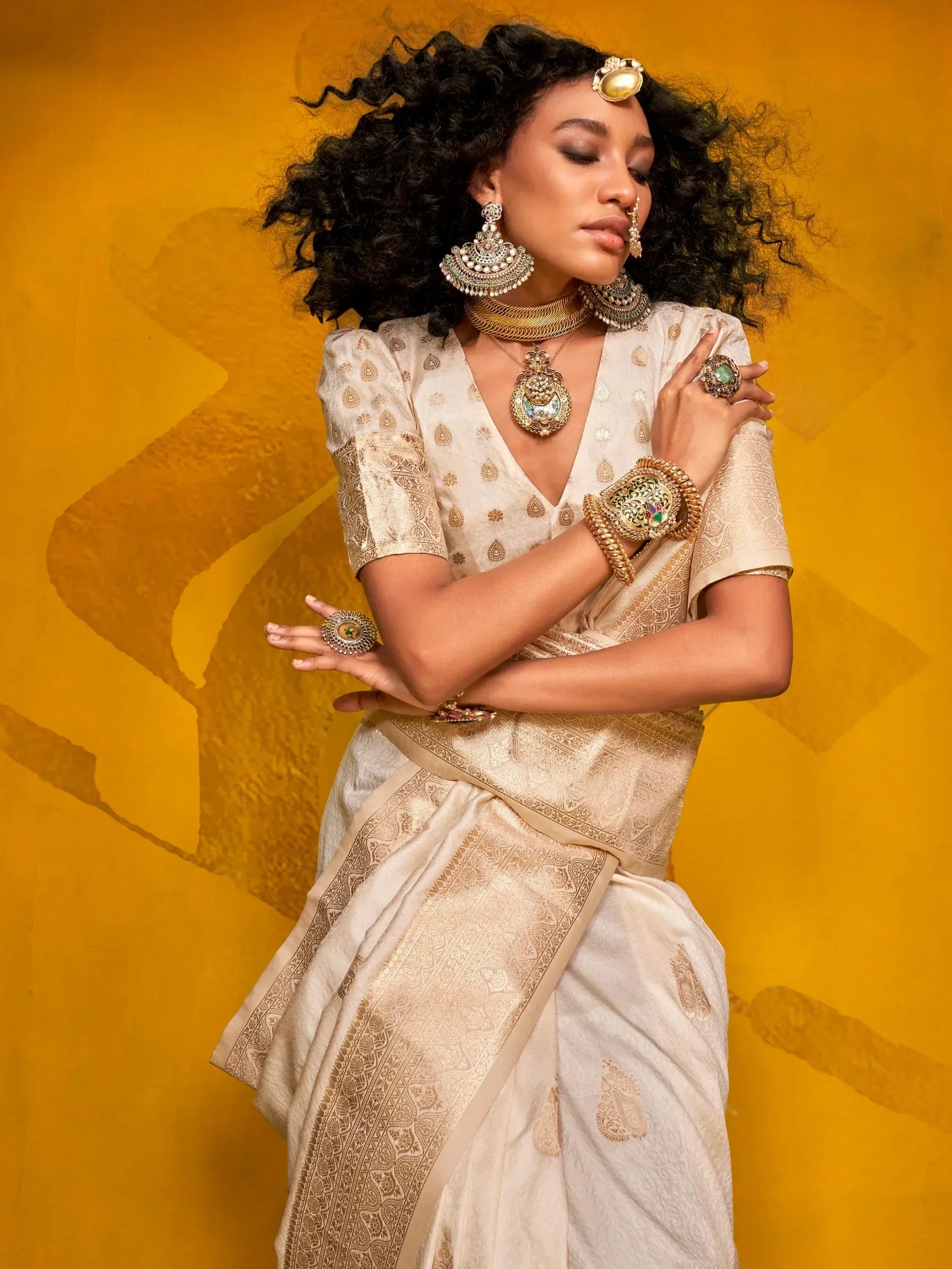 Elegant woman in cream designer saree with gold embroidery, traditional Indian jewelry, against warm yellow backdrop.