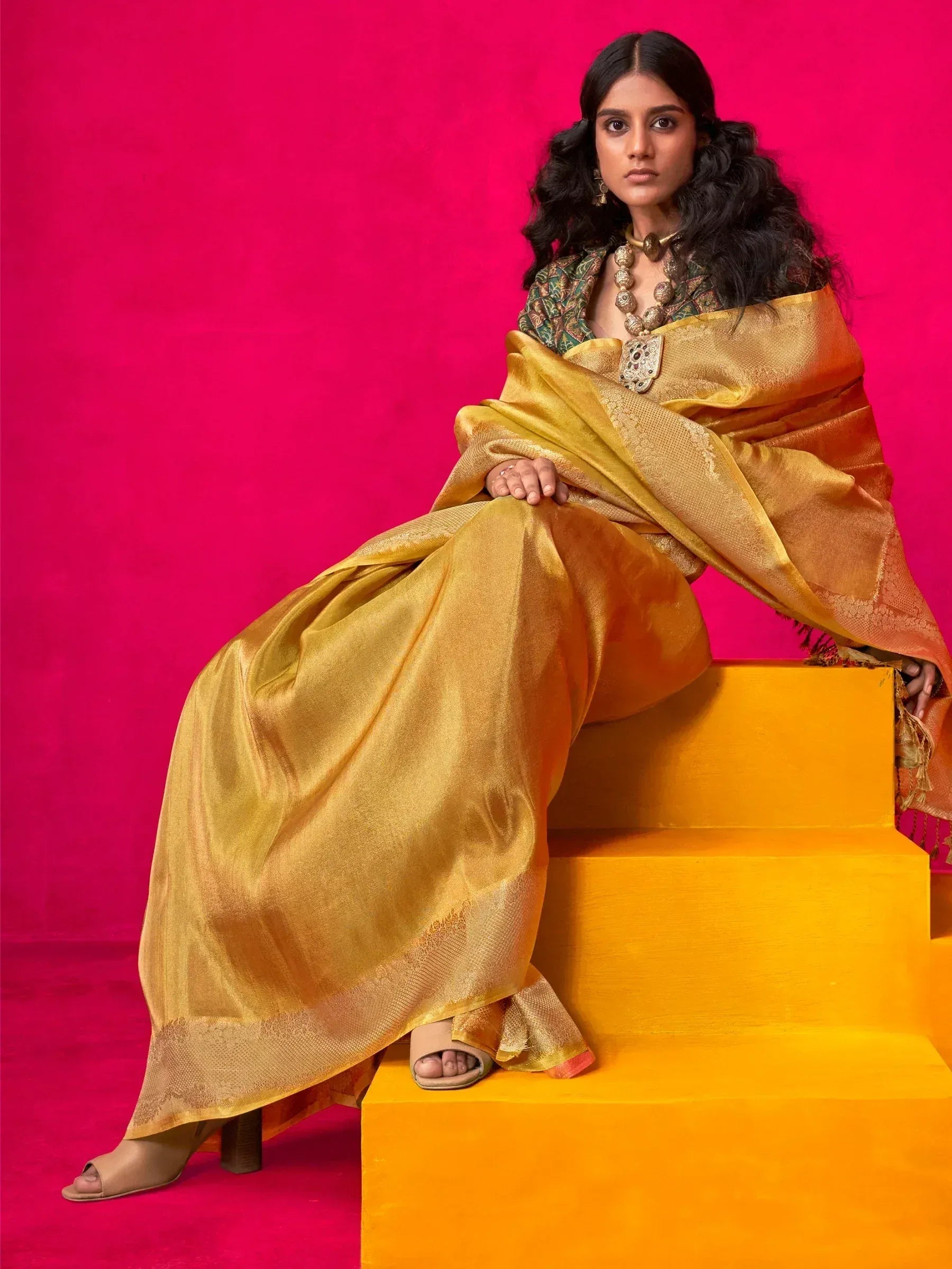 Elegant woman in golden saree with intricate embroidery, styled with traditional jewelry against vibrant pink backdrop.
