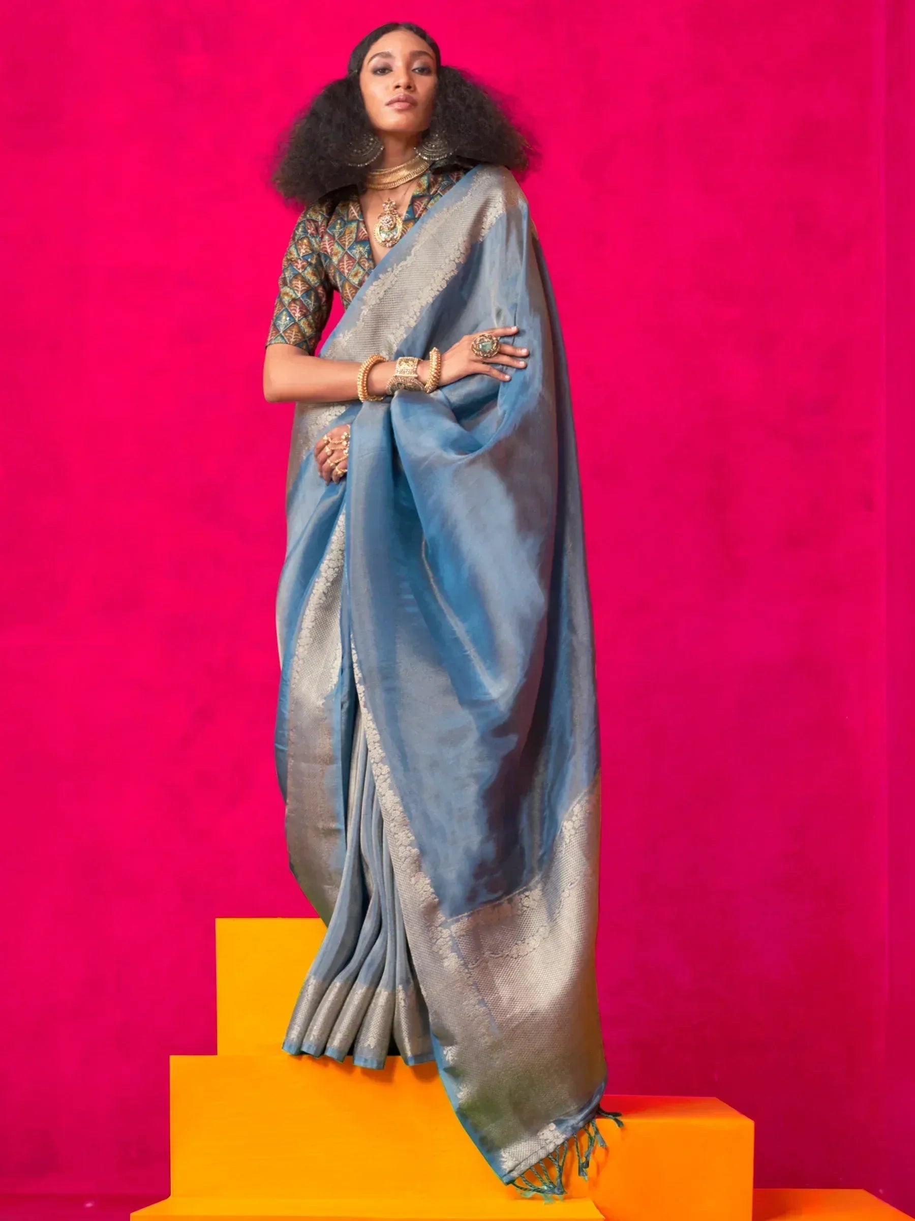 Elegant blue saree with silver border, paired with patterned blouse and gold jewelry, showcased on vibrant pink background.