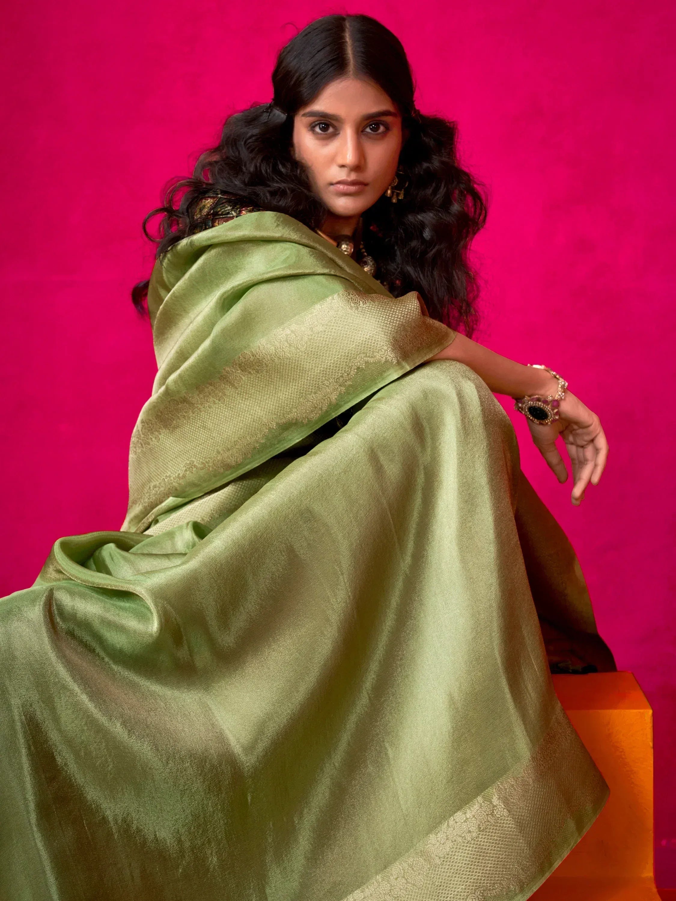 Elegant green saree with golden border, styled with traditional jewelry against a vibrant pink backdrop.