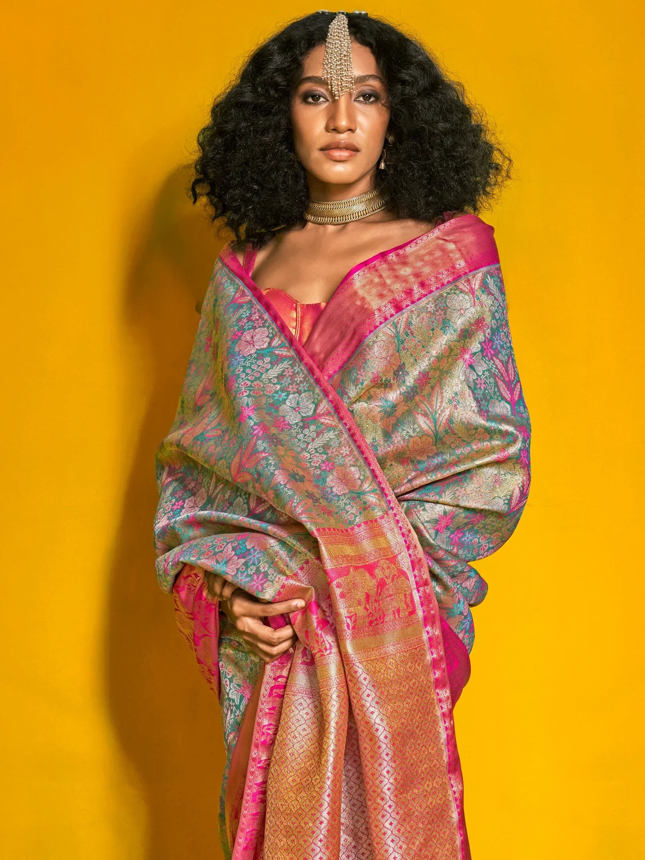 Elegant woman in vibrant floral saree with gold embroidery, styled with traditional jewelry against a yellow backdrop.