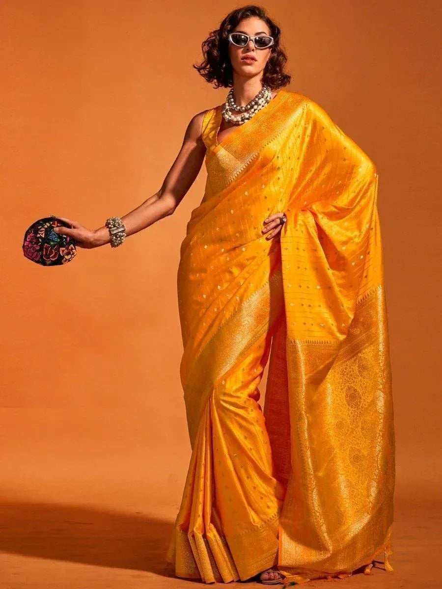 Woman in vibrant yellow silk saree with gold embroidery, styled with sunglasses and floral clutch for ethnic wear
