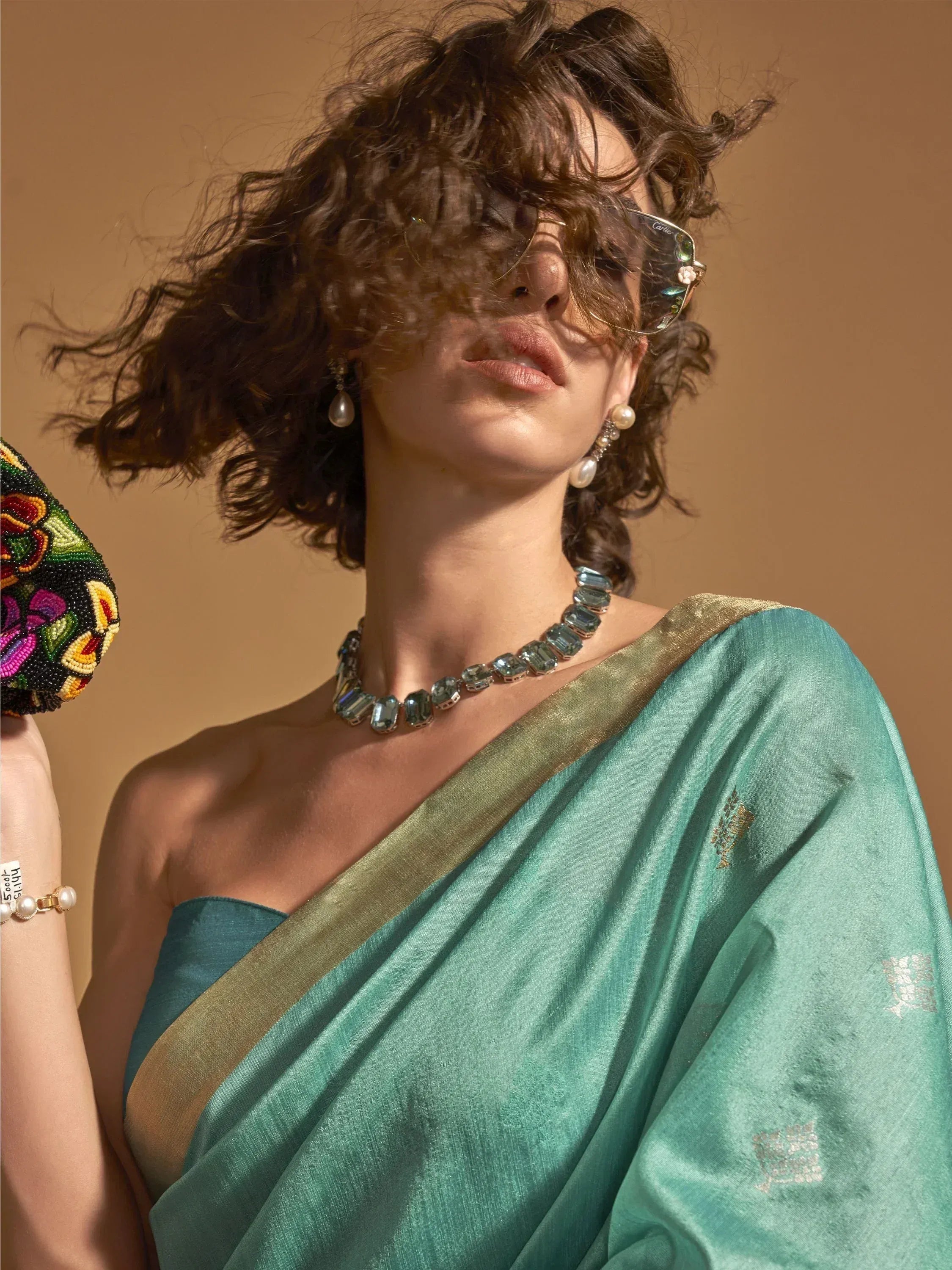 Woman in elegant teal saree with gold border, statement necklace, and pearl earrings, styled with sunglasses.