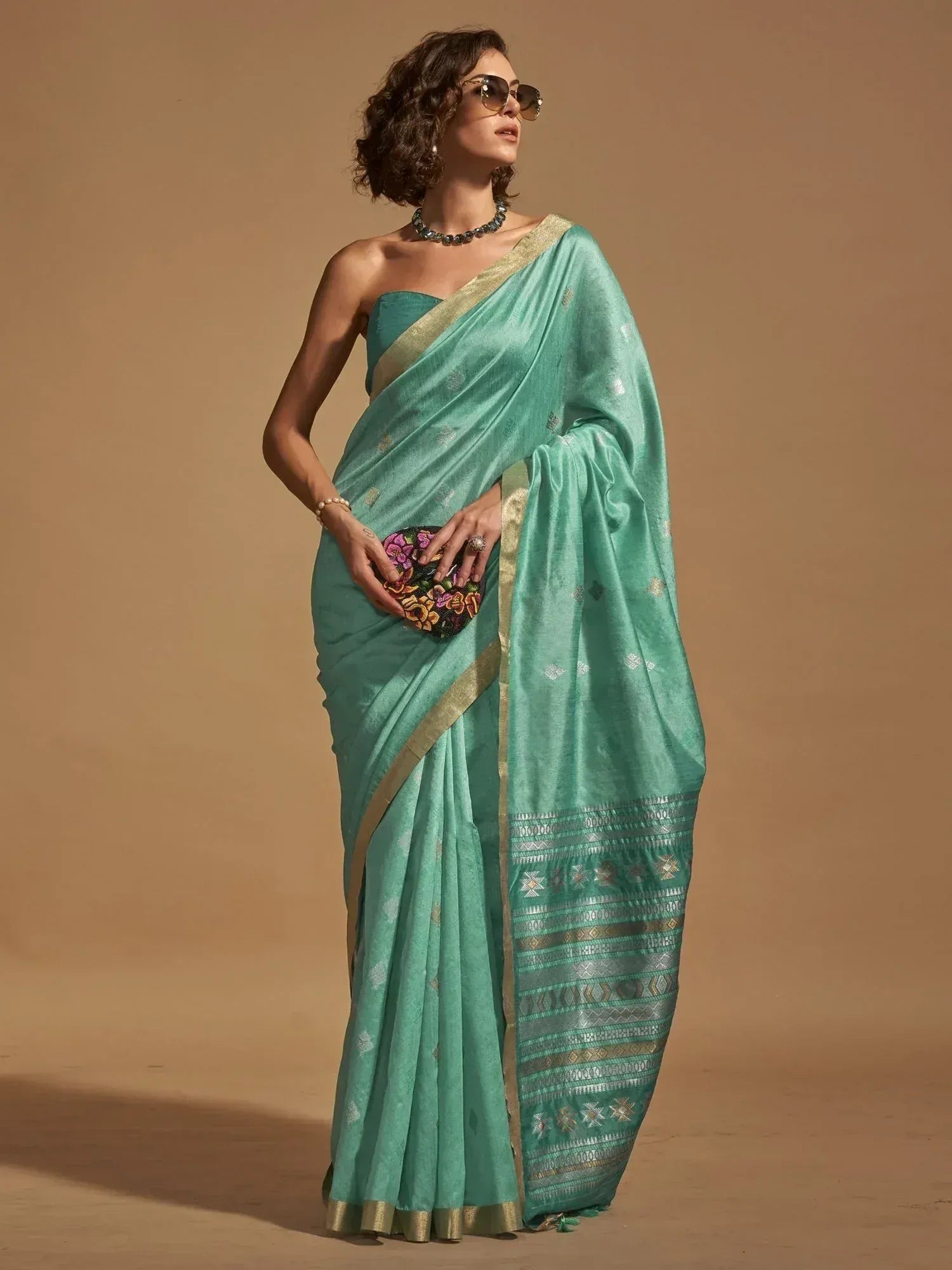 Teal designer saree with gold border and ethnic print, paired with floral clutch and sunglasses