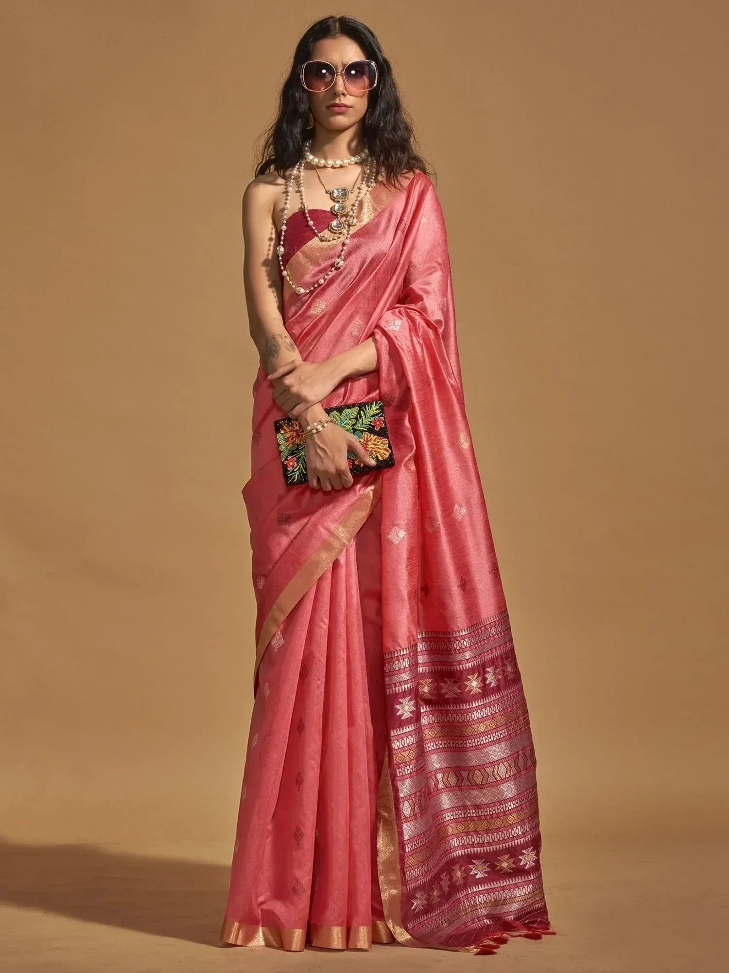 Pink designer saree with gold border and geometric patterns, paired with pearl jewelry and floral clutch