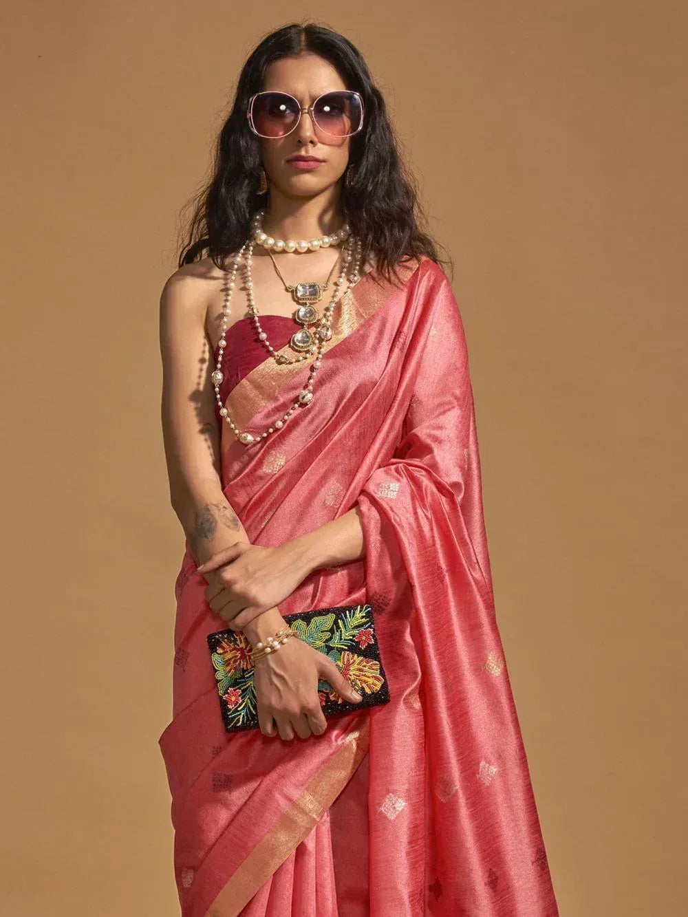 Woman in pink designer saree with gold border, pearl jewelry, and floral clutch, styled for ethnic occasions
