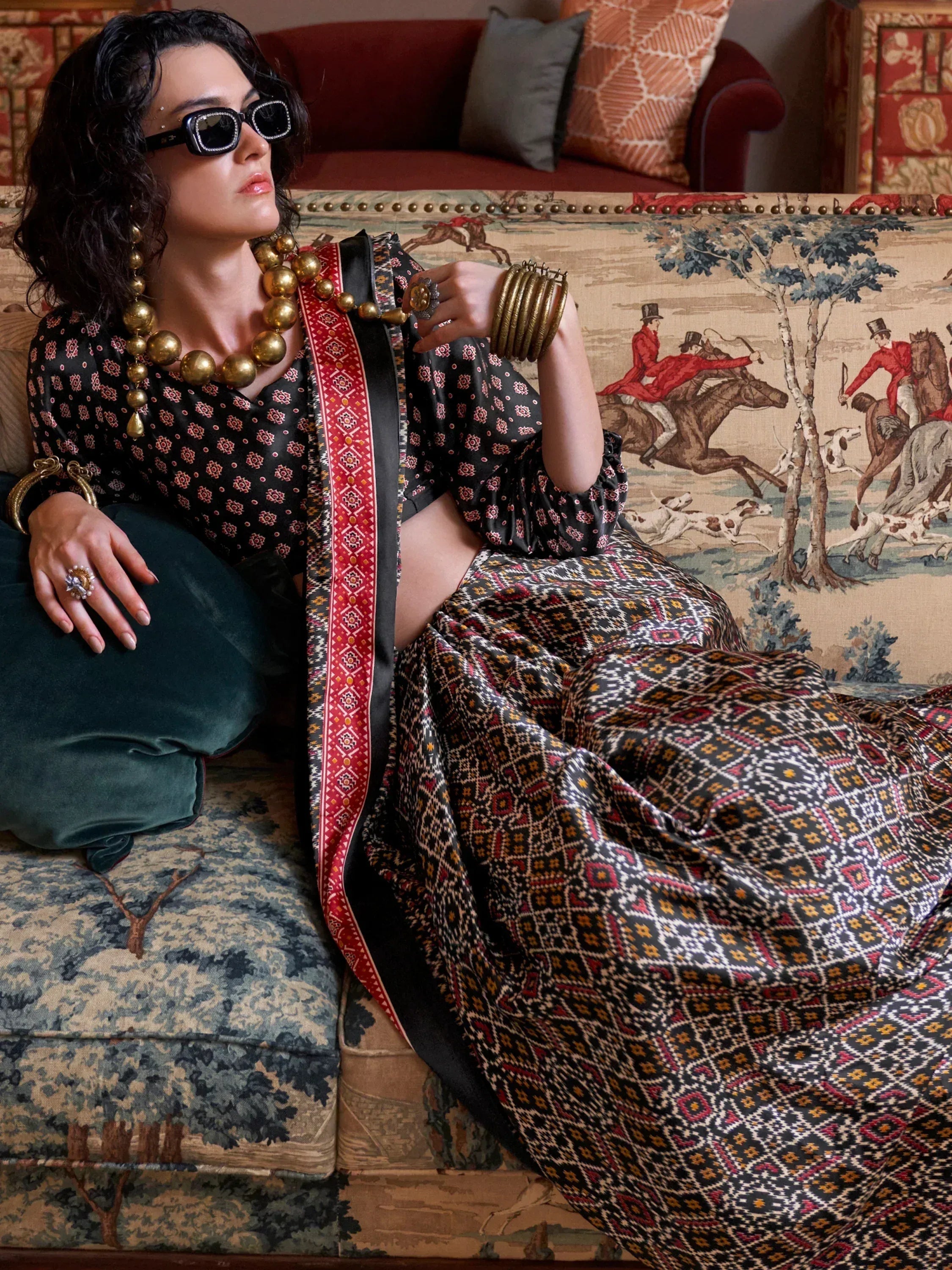 Woman in ethnic print gown with gold jewelry, styled on patterned sofa for Indian fashion look