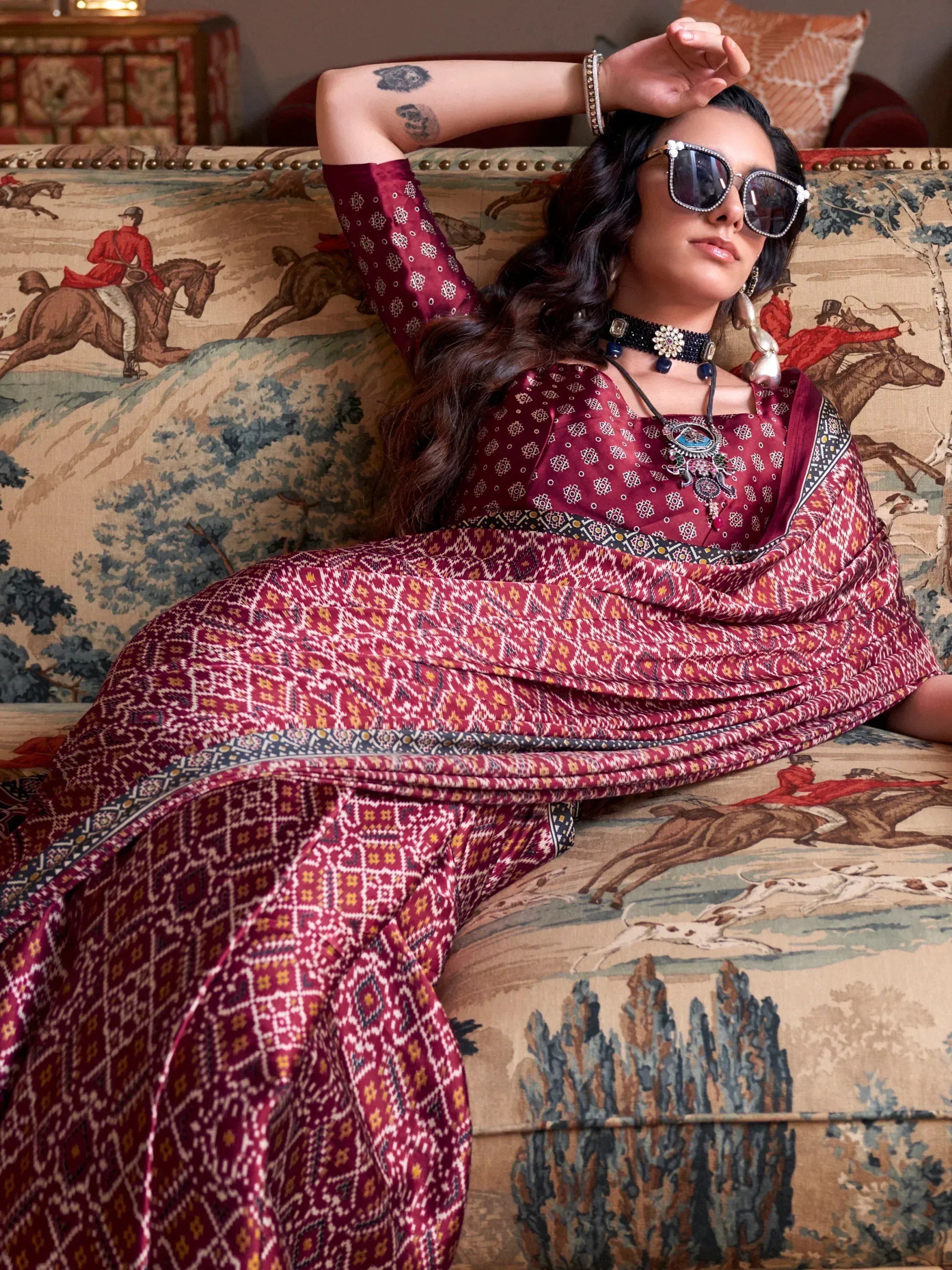 Woman in maroon printed ethnic gown with sunglasses, styled on patterned sofa, perfect for Indian festive wear