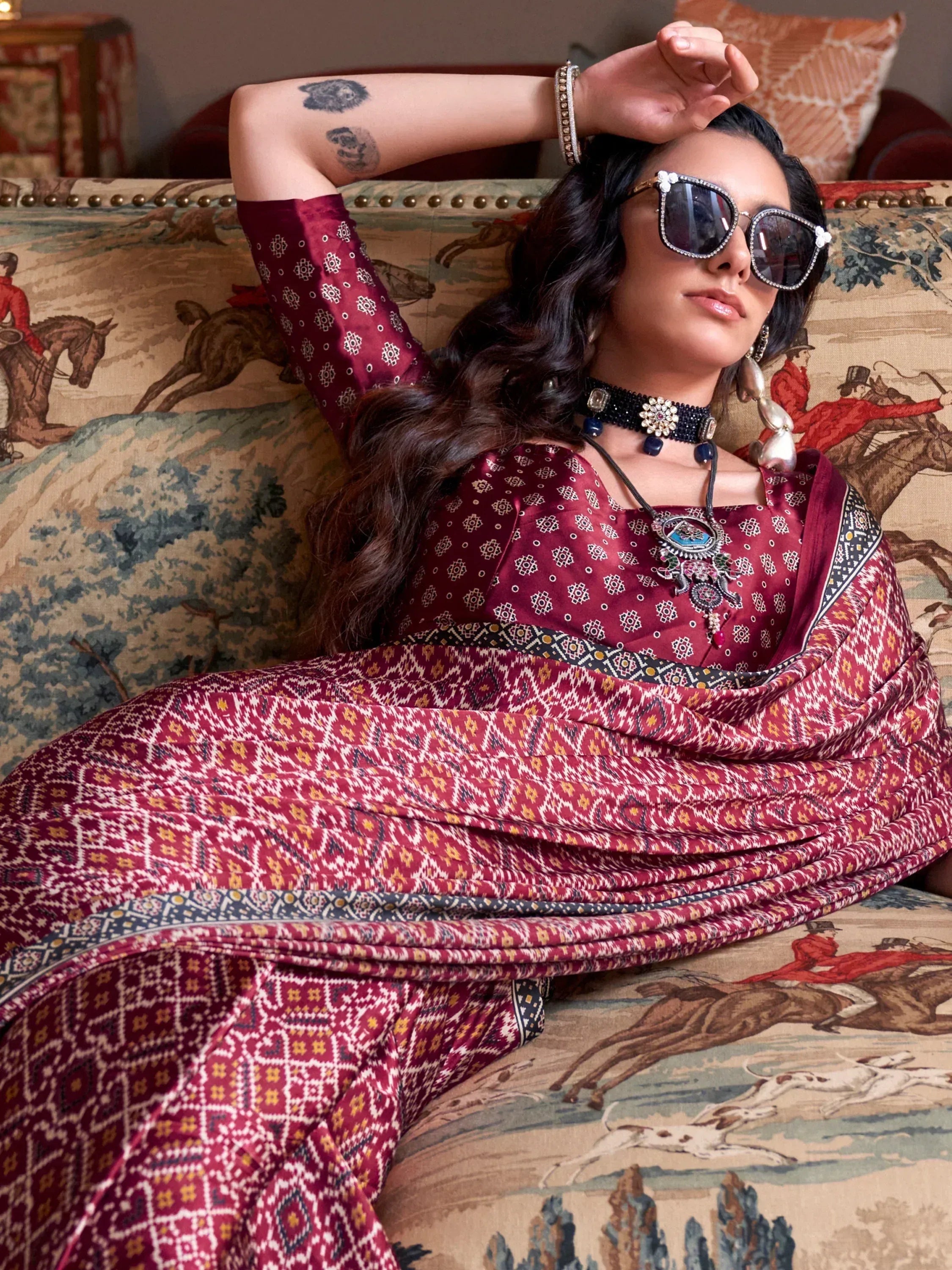 Woman in maroon ethnic salwar kameez with geometric print, styled with sunglasses and jewelry, lounging on patterned sofa