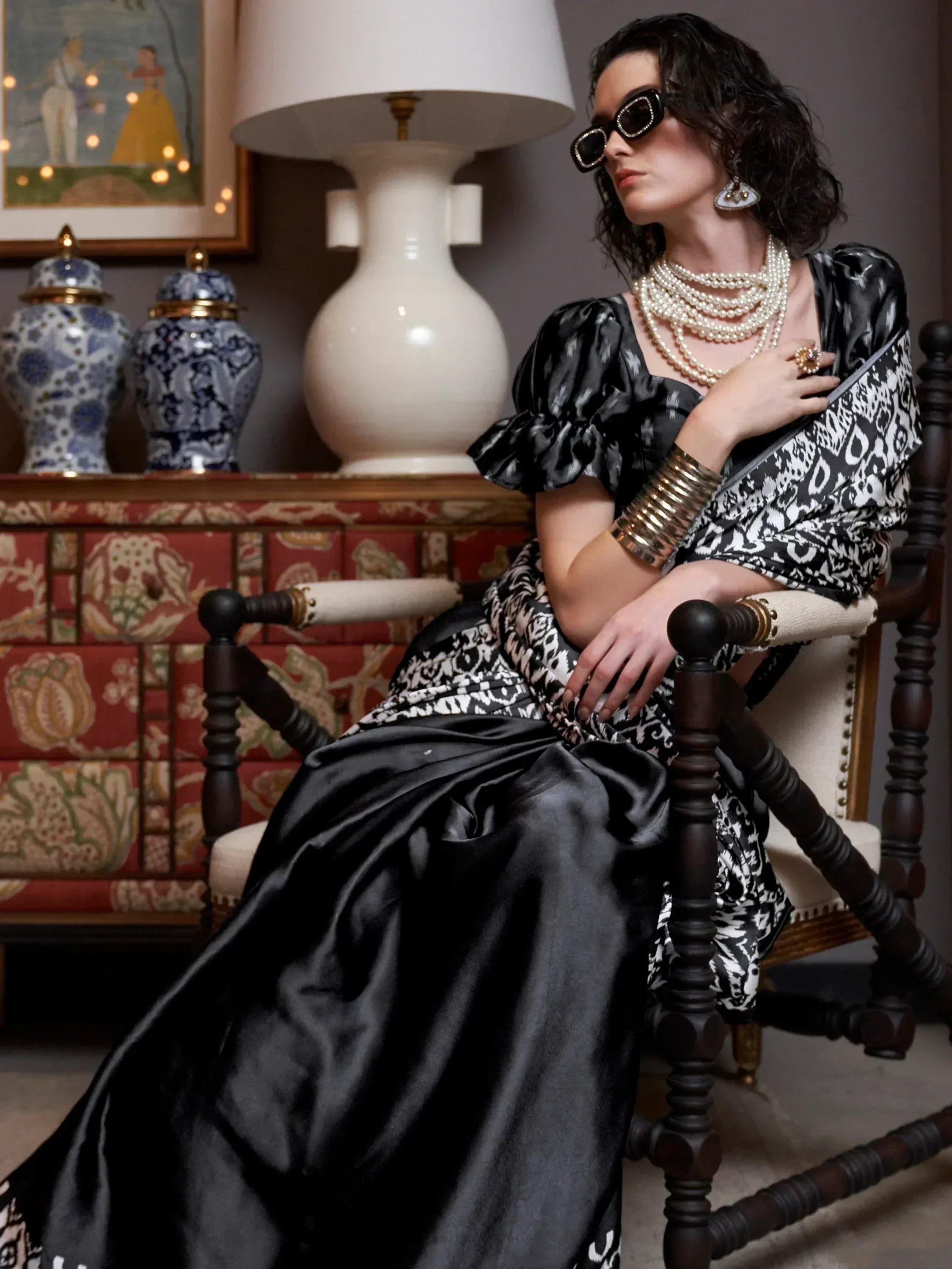 Woman in black and white ikat print ethnic gown with pearl jewelry, seated elegantly in vintage chair