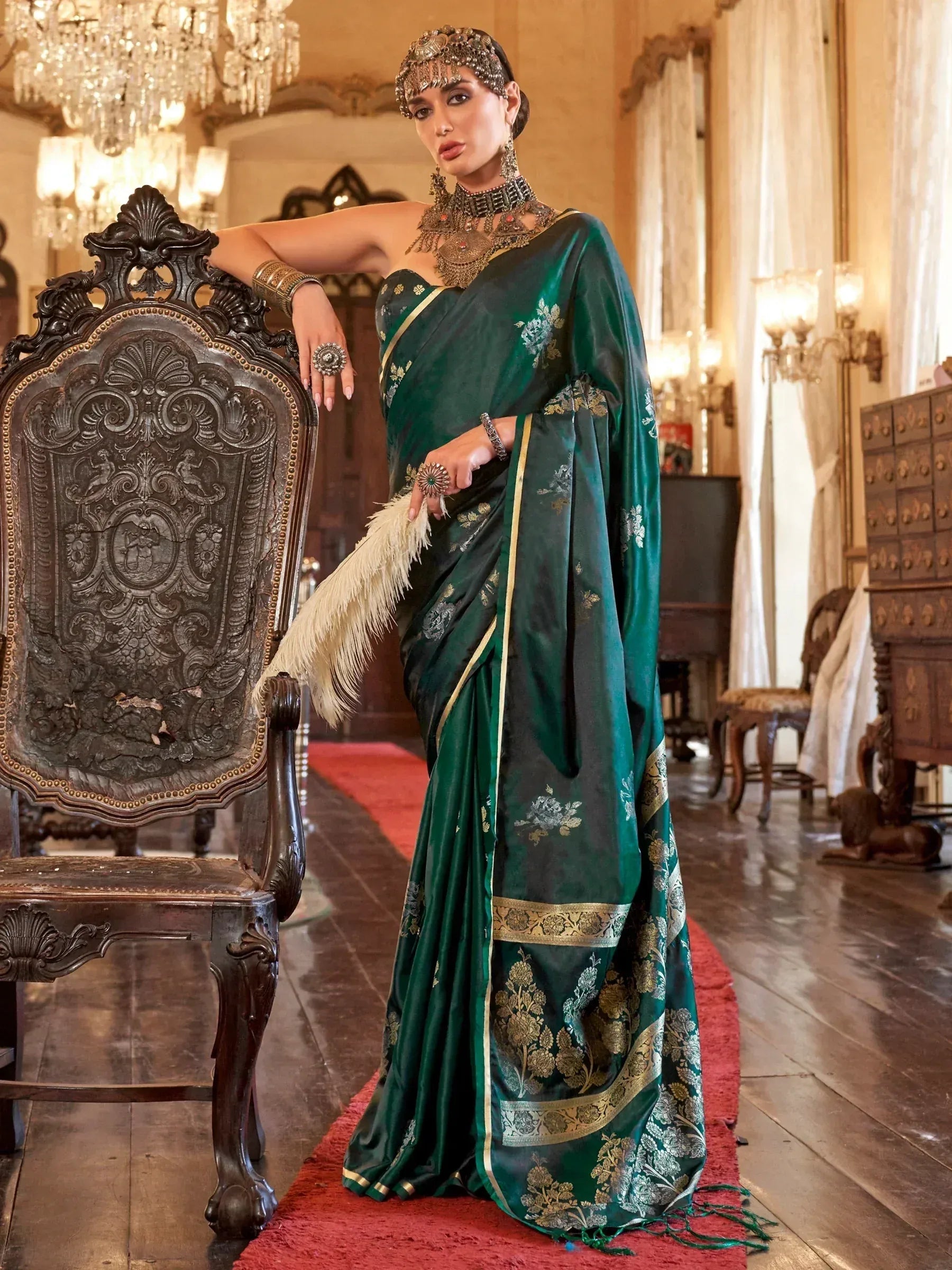 Elegant woman in green designer saree with gold embroidery, traditional jewelry, and feather fan, posing beside ornate chair.